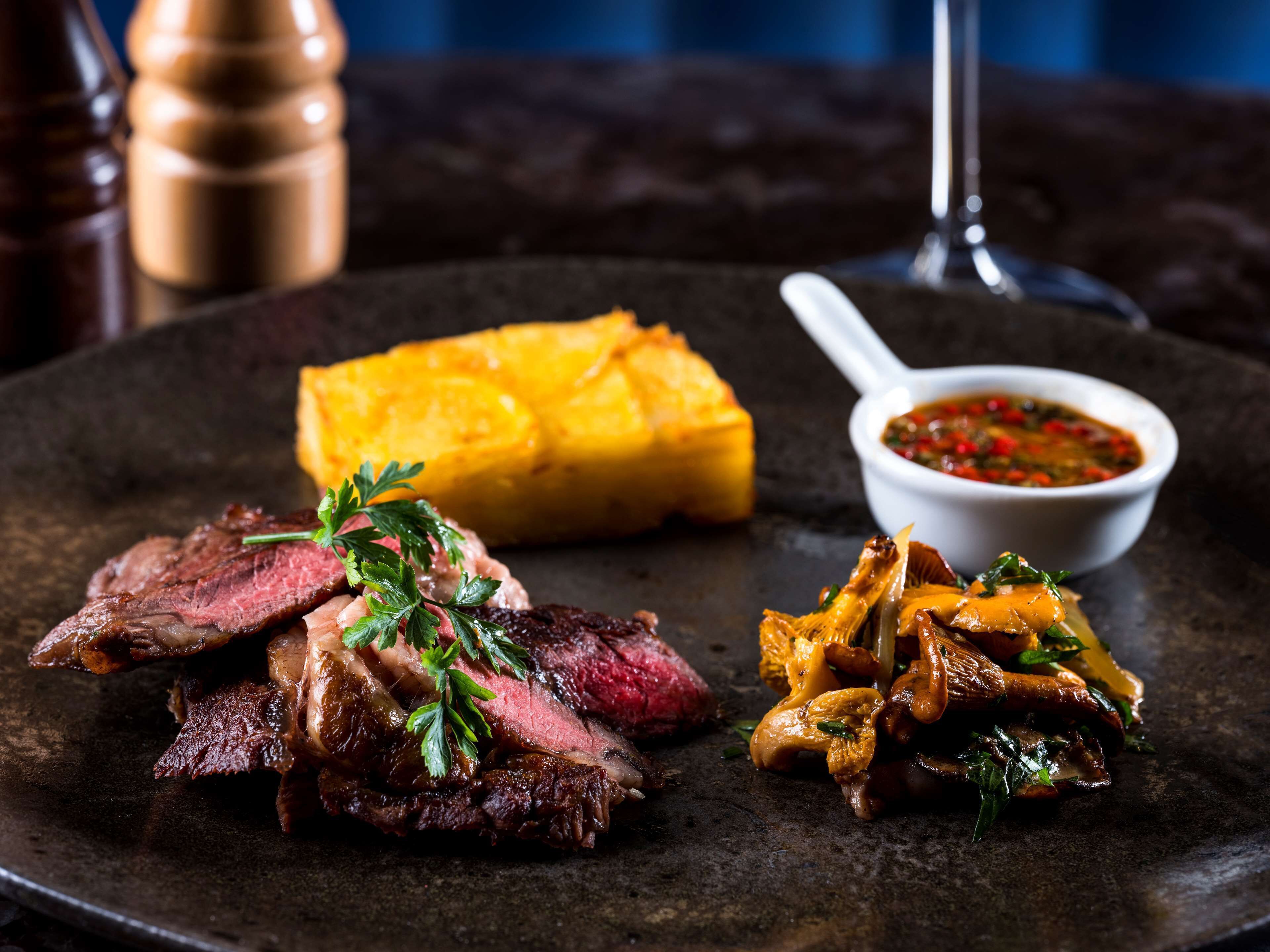 A plate of sliced steak with a garnish, a piece of yellow squash, and a bowl of red sauce.