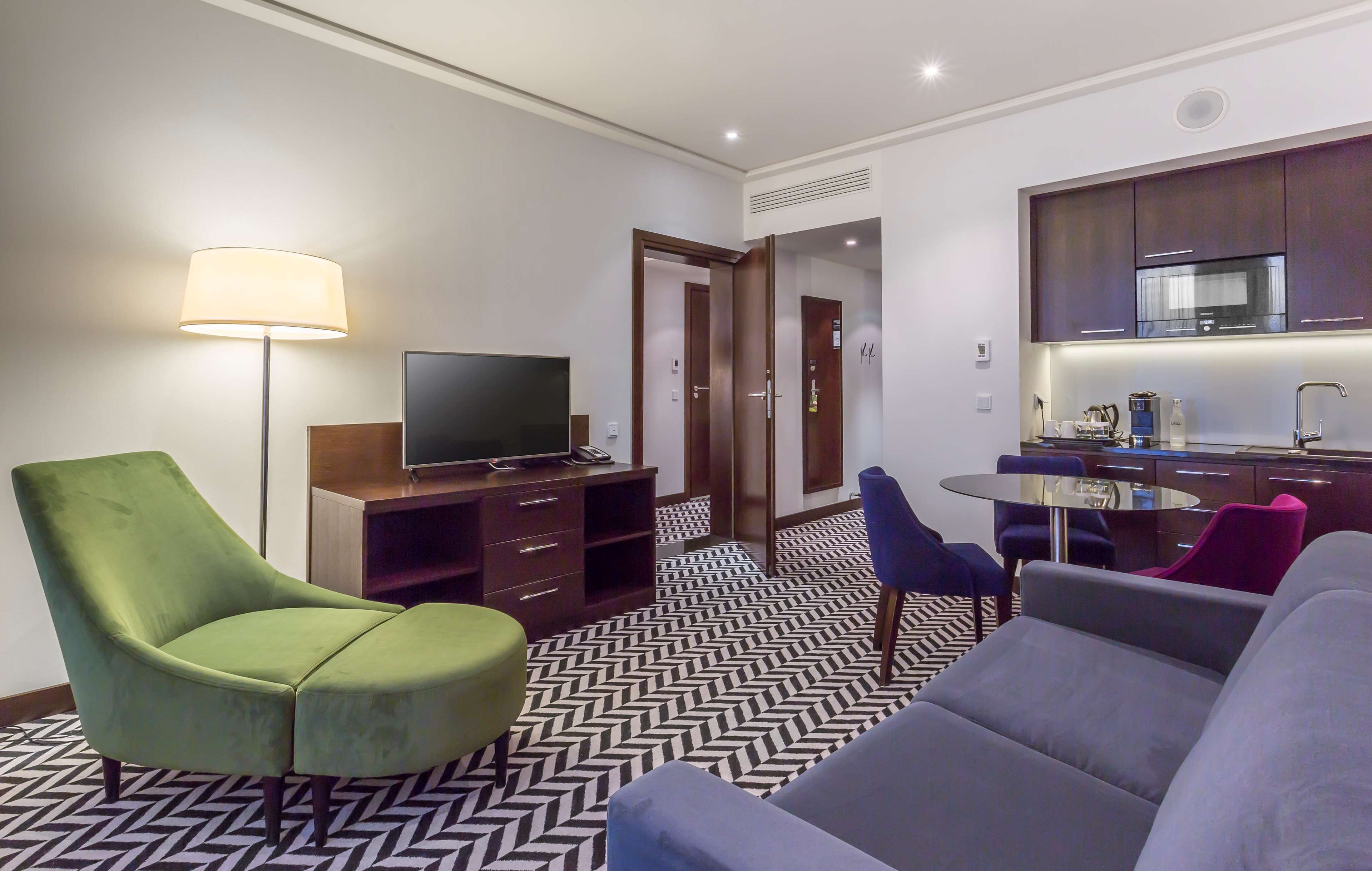A modern hotel room with a sofa, armchair, flat-screen TV, and a small dining table.