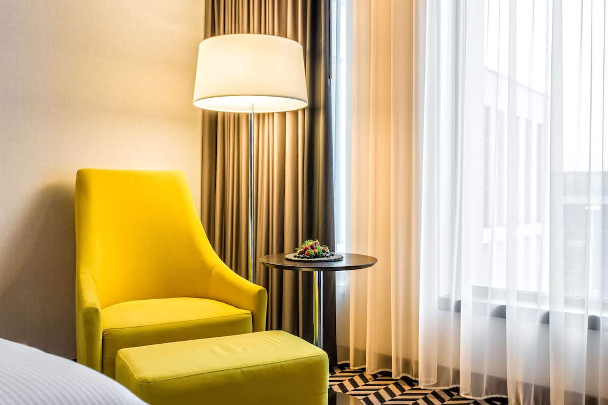 Une chambre d’hôtel avec un fauteuil jaune, une petite table ronde ornée d’une plante et une grande fenêtre avec des rideaux légers.