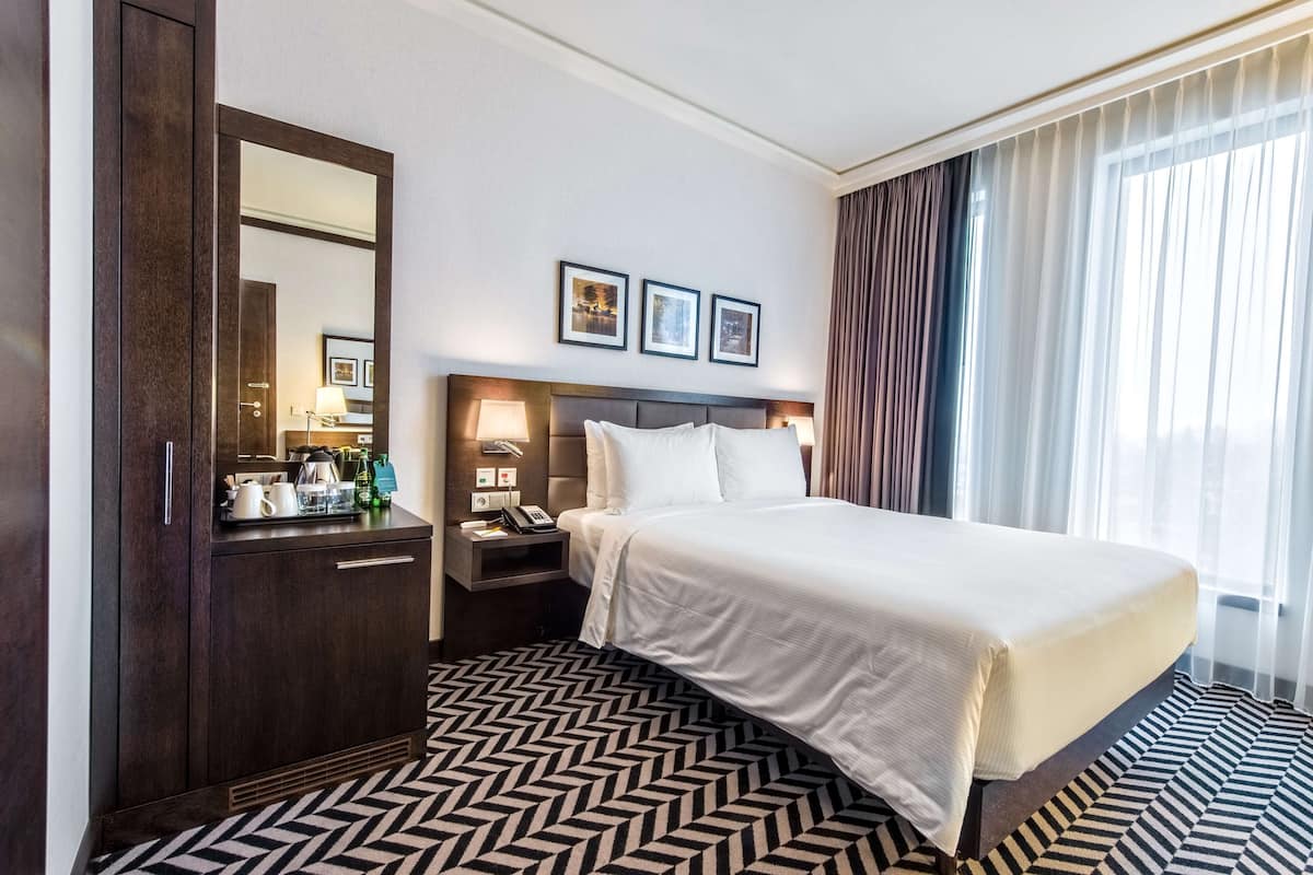 Une chambre d’hôtel avec un grand lit, une commode, une table de chevet, un miroir et une fenêtre avec des rideaux.