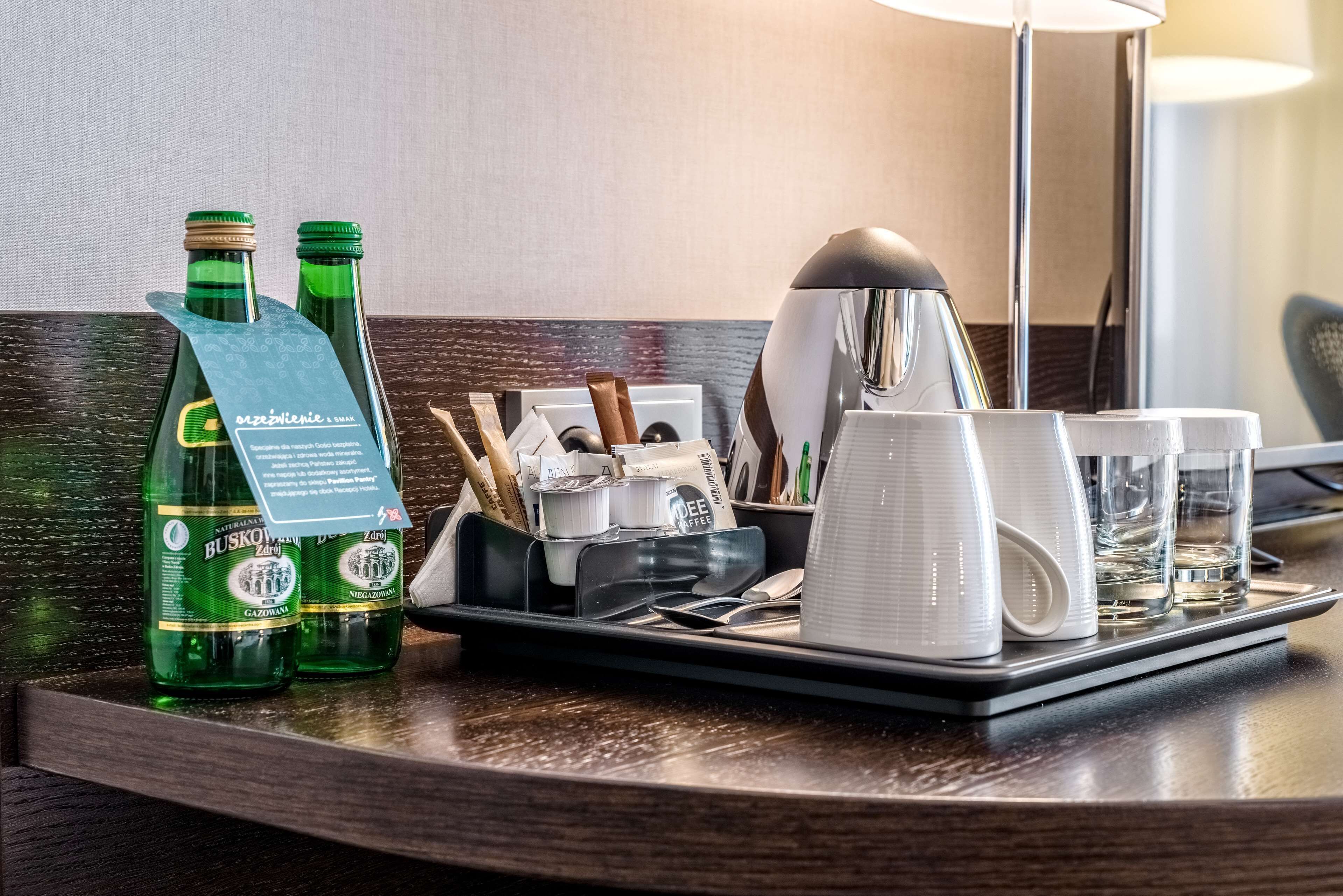 A hotel minibar setup with green bottles, a coffee machine, and white cups and glasses.
