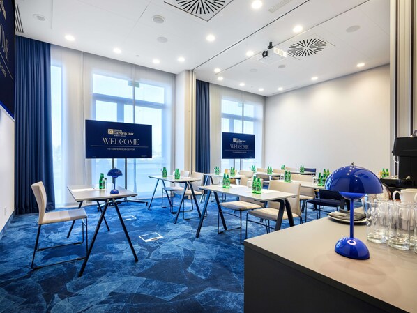 A conference room with a blue carpet, white tables, and chairs, equipped with water bottles and a coffee machine.