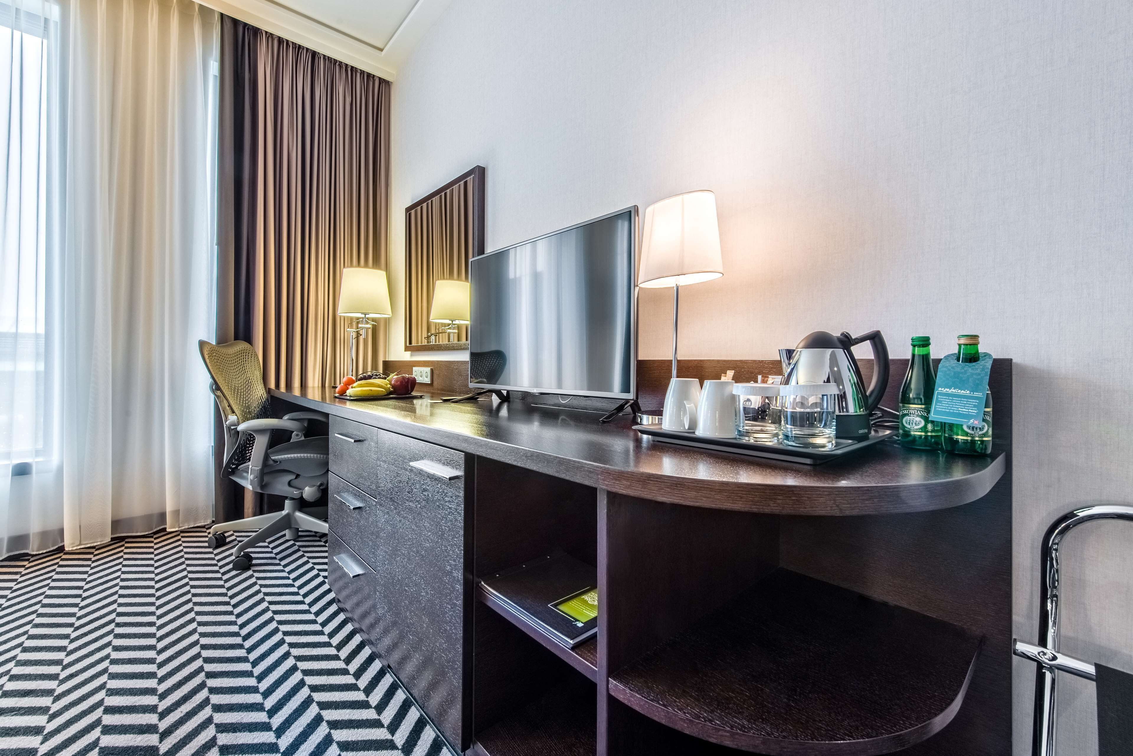 Habitación de hotel con escritorio, silla, televisor, lámpara, cafetera y bandeja con botellas y vasos.