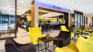A modern lounge area with yellow and black chairs, a bar with a visible name sign, and a glass wall offering a view of the city.