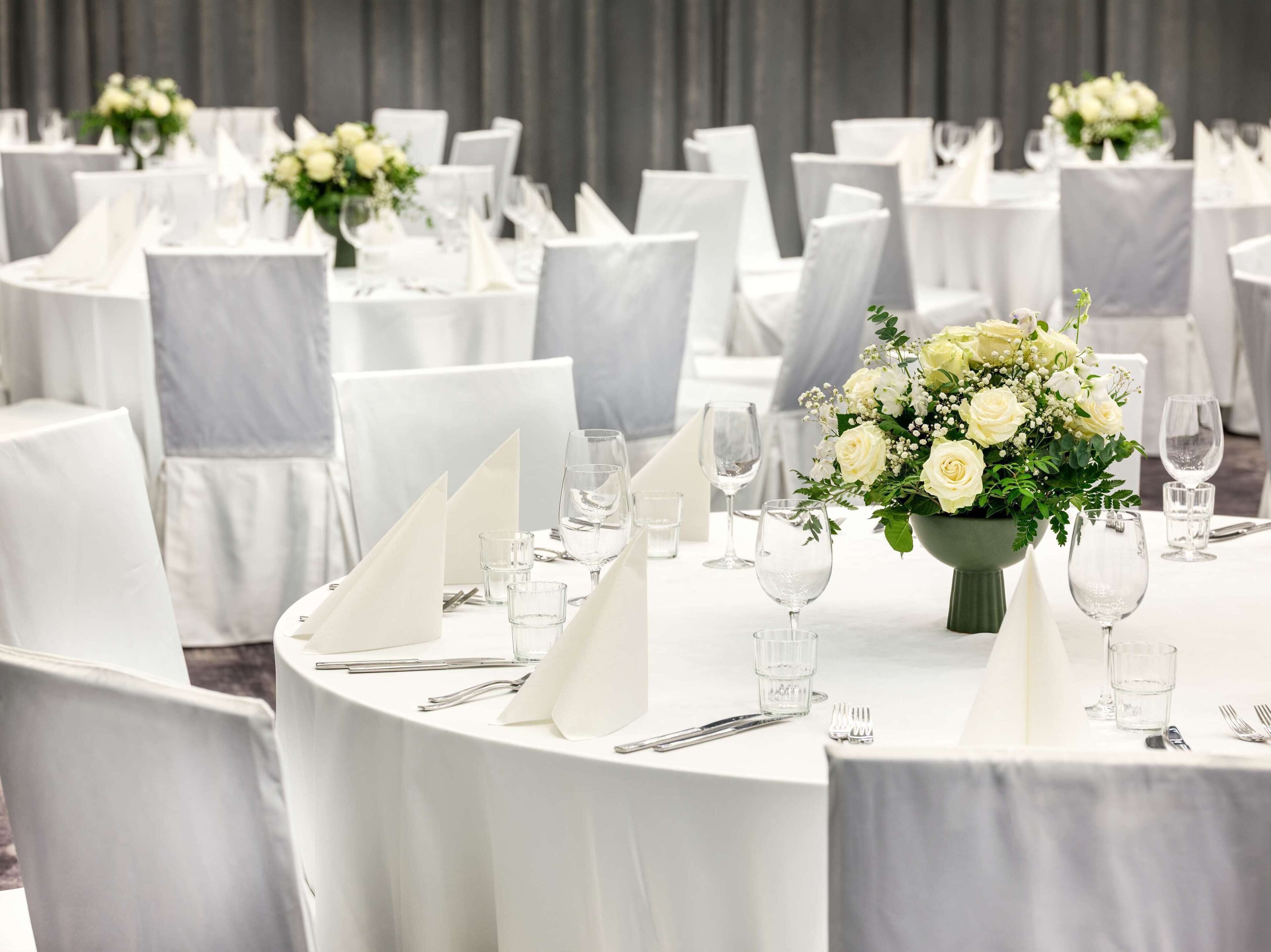 A banquet hall with round tables, white tablecloths, and matching chairs. Each table is set with cutlery, glasses, and a centerpiece of white flowers.