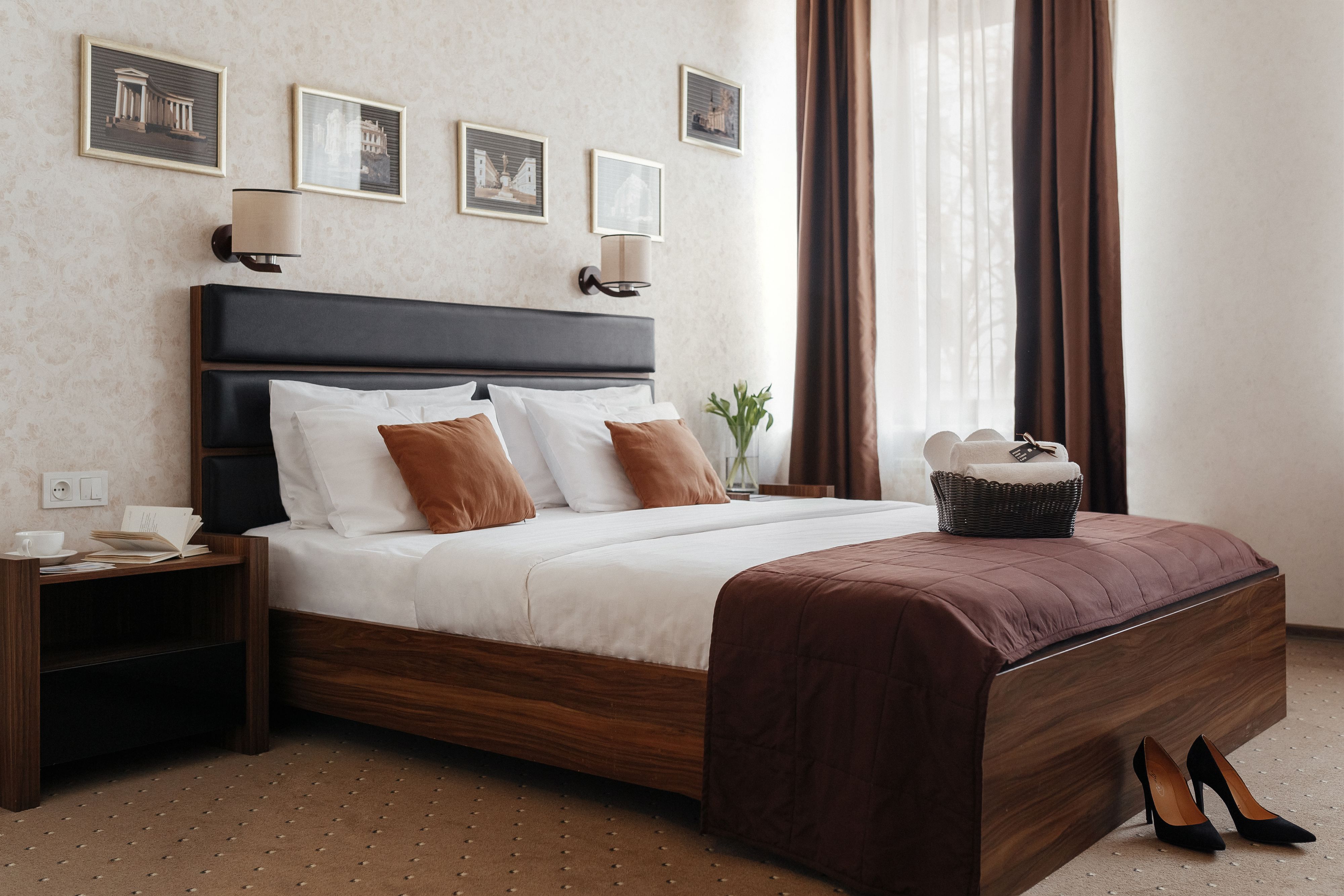 A neatly made bed with a dark headboard, white and brown pillows, and a brown throw blanket. There are framed pictures on the wall above the bed, a nightstand with a lamp, and a basket with a fork and knife on the side.