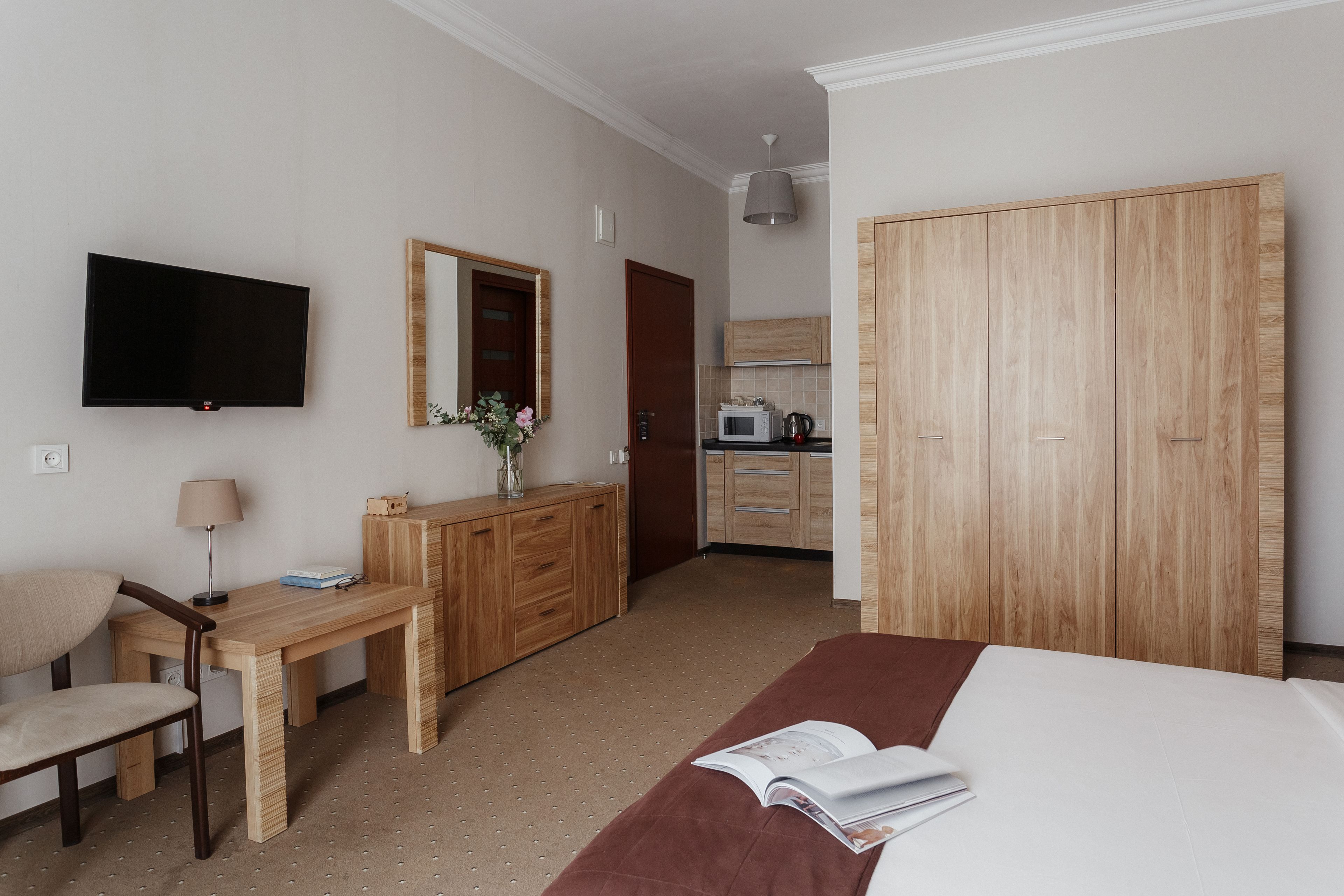 A hotel room with a bed, a desk, a chair, a TV, a mirror, a wardrobe, and a kitchenette.
