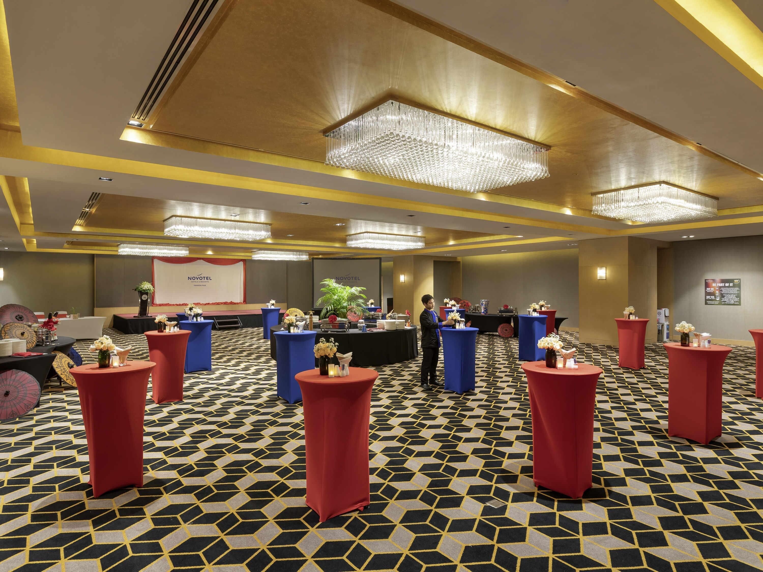 A hotel lobby with a check-in counter, red and blue round tables, and a patterned carpet.