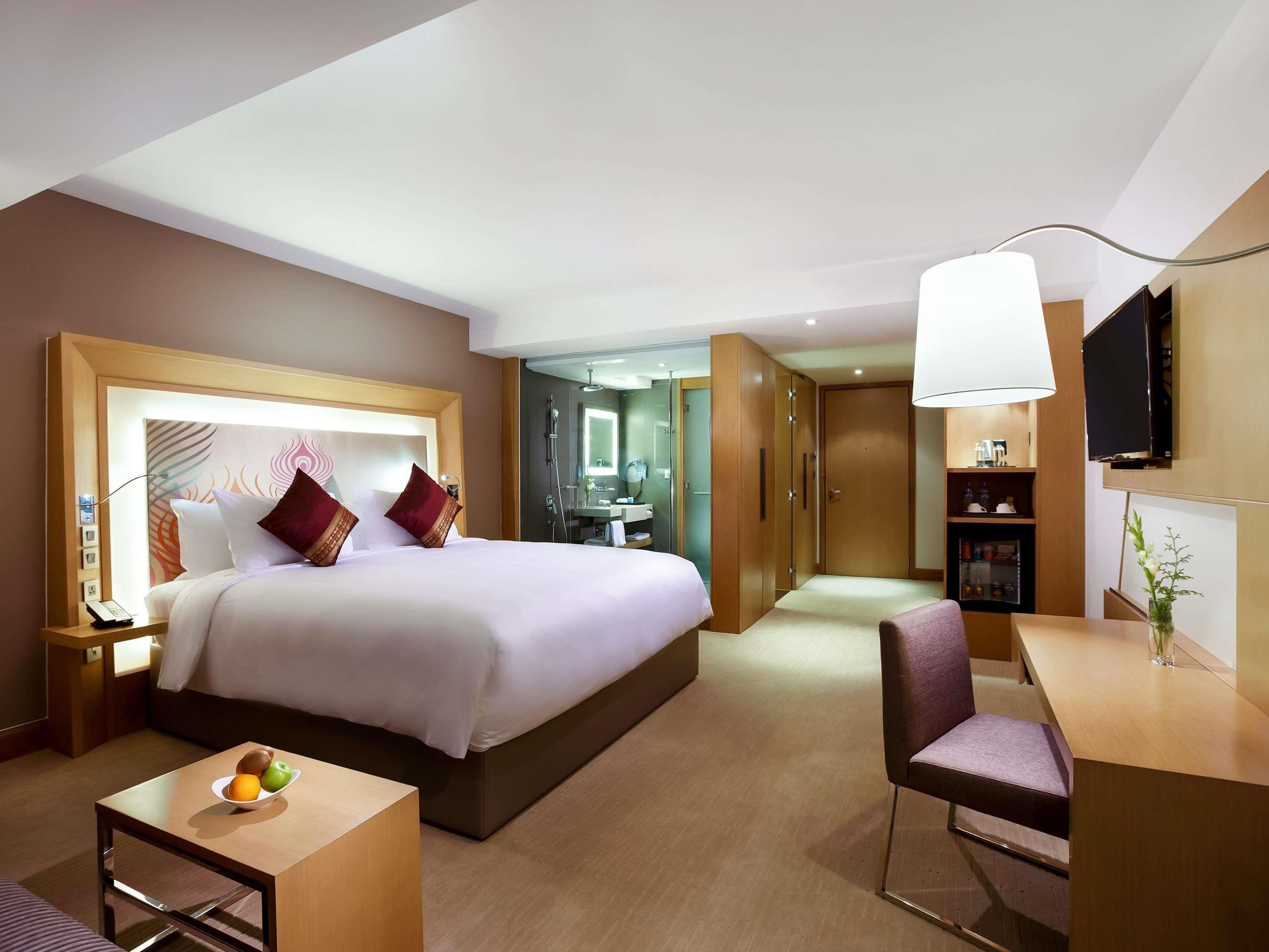 A hotel room with a large bed, a desk, a chair, a small table with fruit, a mirror, a wardrobe, and a TV on the wall.