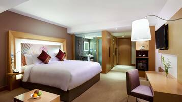 A hotel room with a large bed, a desk, a chair, a small table with fruit, a mirror, a wardrobe, and a TV on the wall.