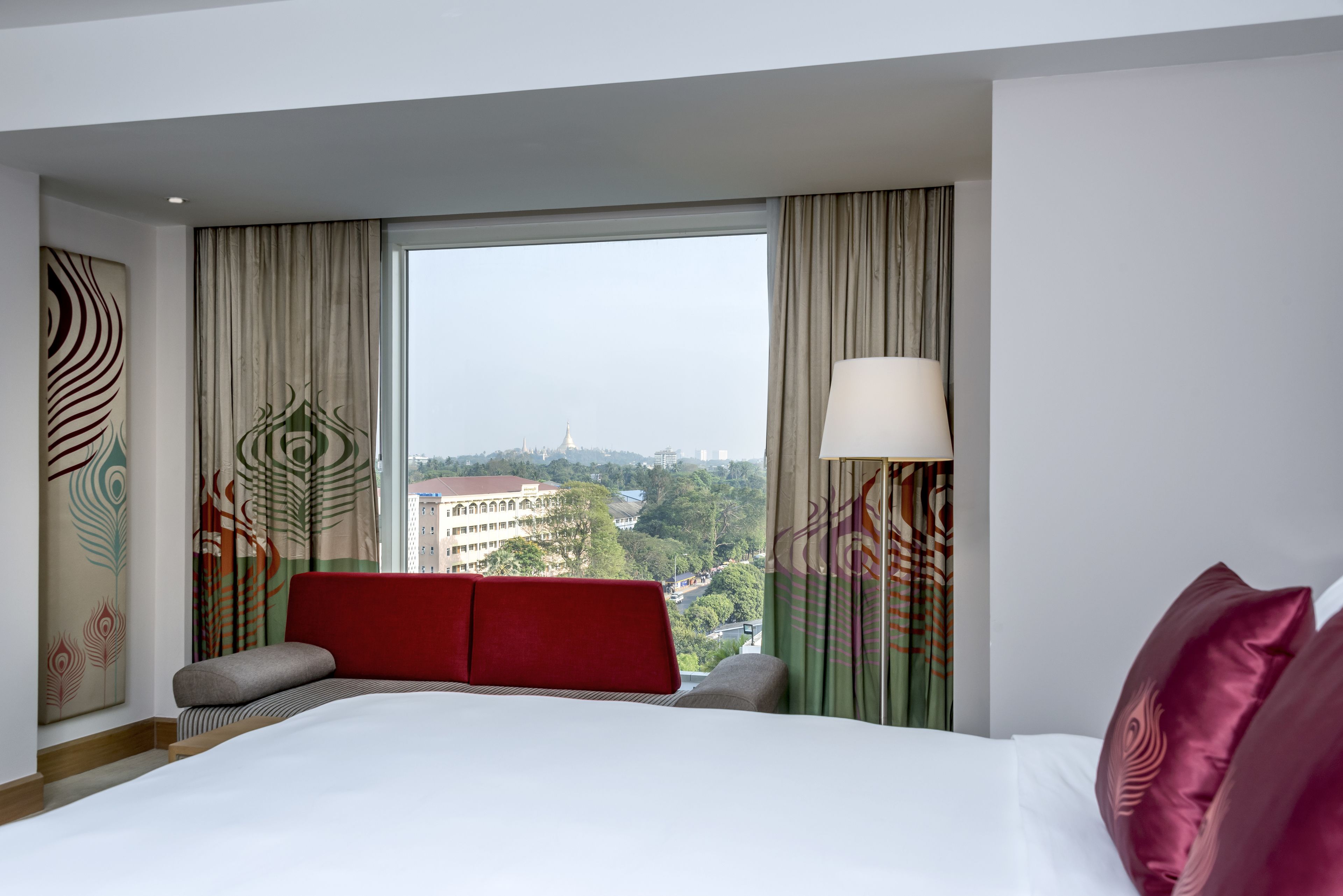Habitación de hotel con una cama grande, un cabecero rojo, vista de un paisaje urbano a través de un ventanal amplio y un adorno en la pared.
