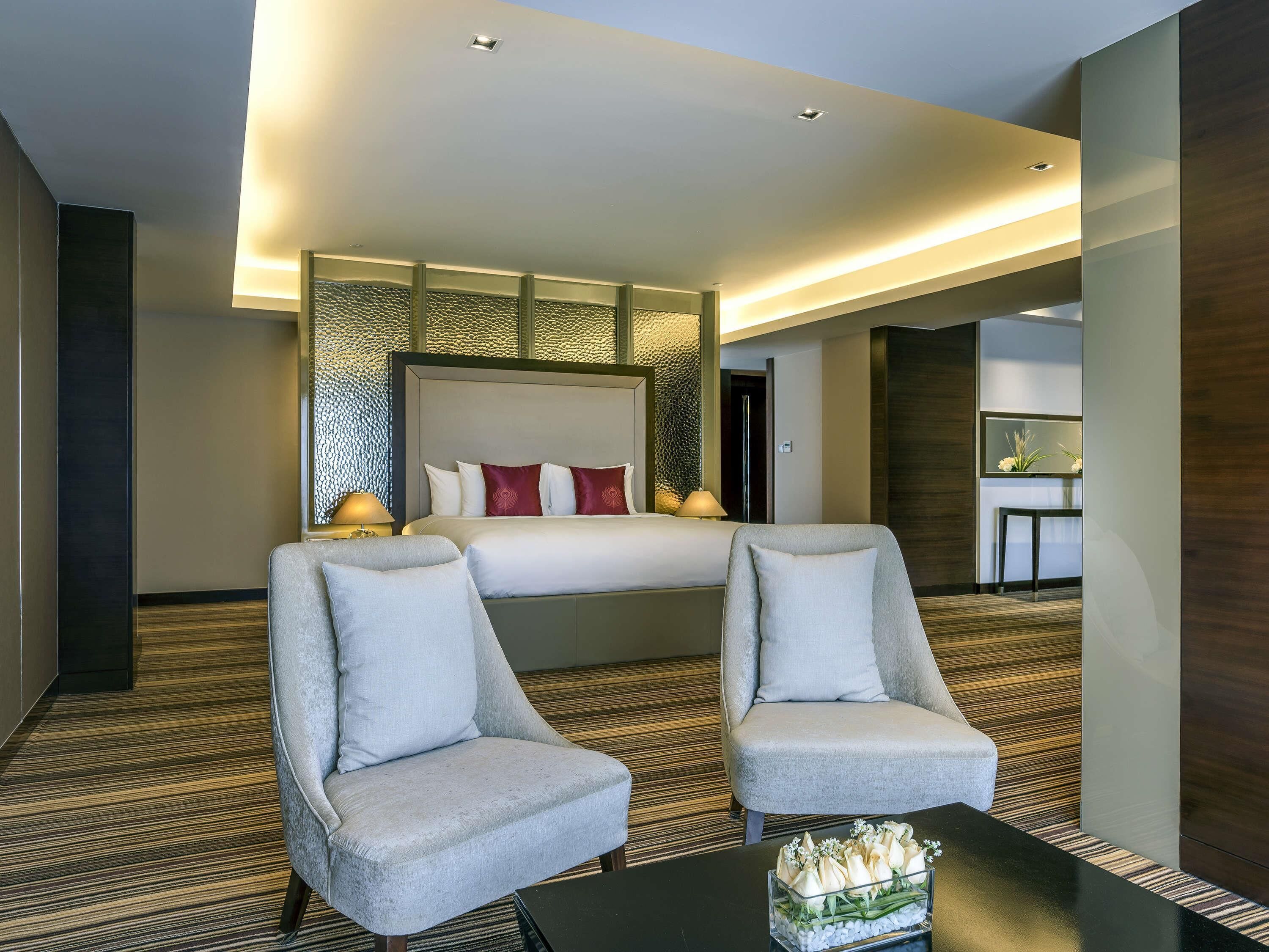 Una habitación de hotel con una cama grande, dos sillones, una mesita con un jarrón de flores y un espejo en la pared.