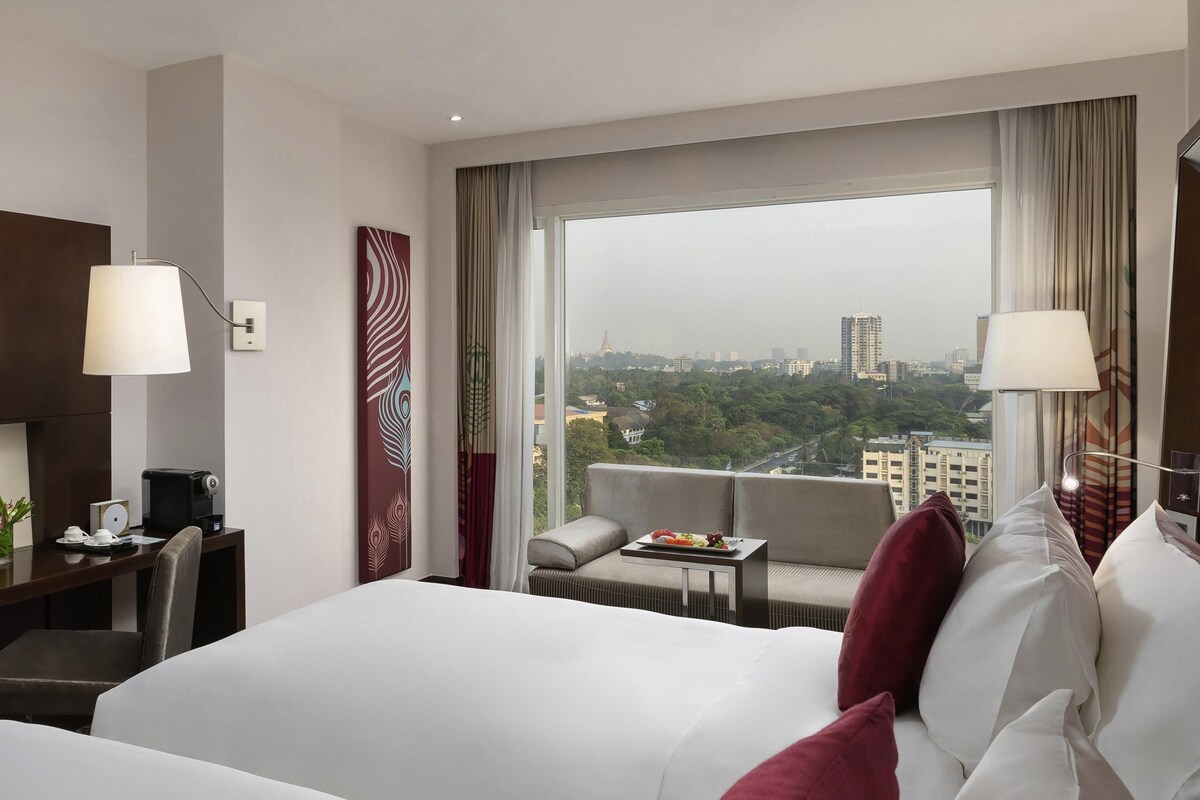 Habitación de hotel con cama, escritorio, silla, una mesita con bandeja de frutas y vistas a la ciudad a través de amplias ventanas.