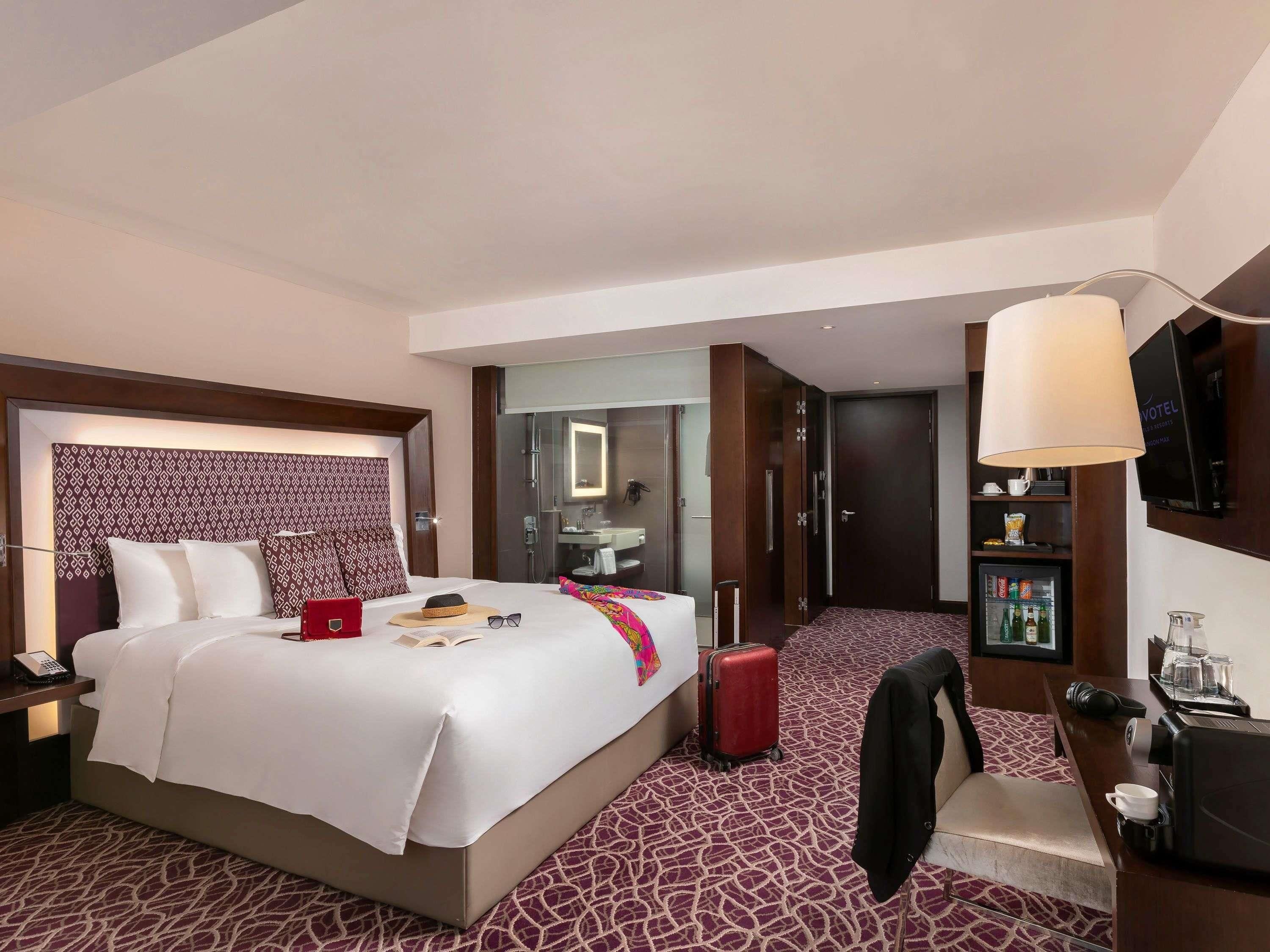 A hotel room with a bed, a desk, a chair, a suitcase, a television, and a minibar.