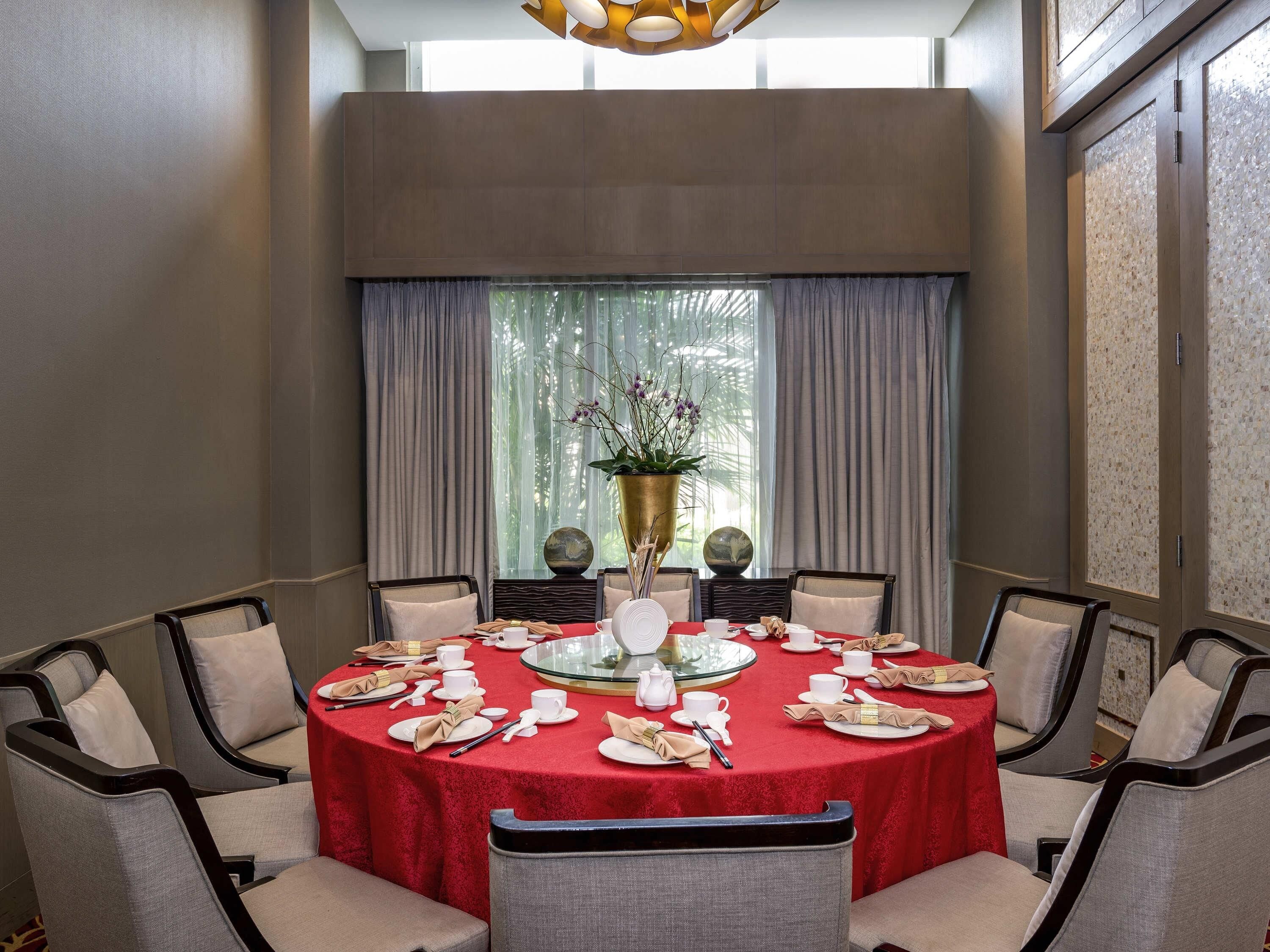 Una mesa redonda con mantel rojo, rodeada por seis sillas, en una habitación con ventanas amplias y cortinas.