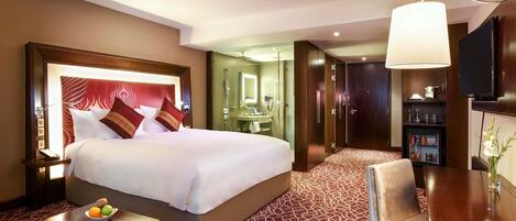 A hotel room with a large bed, a bedside table with fruit, a seating area with a chair, a desk with a lamp, and a wardrobe.