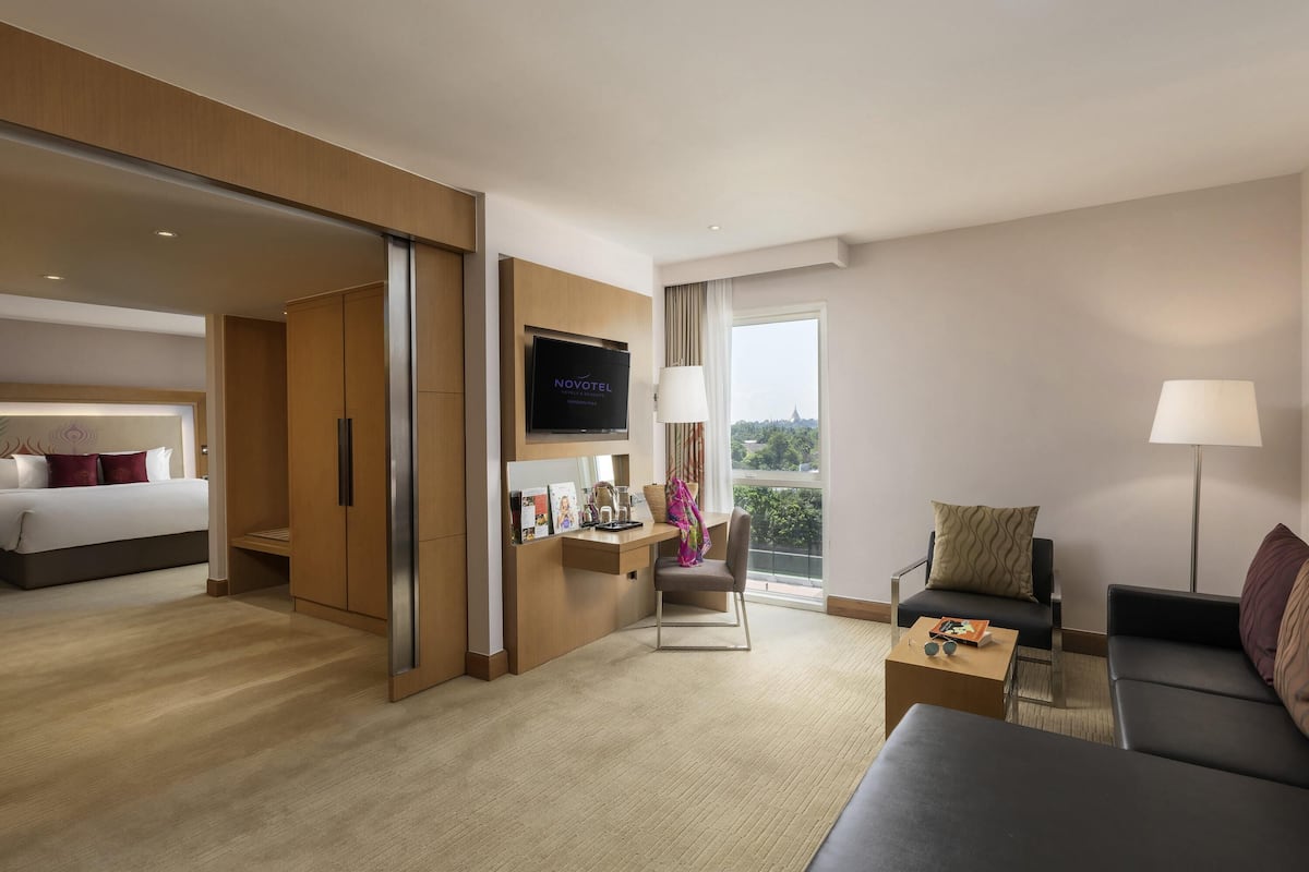 A hotel room with a bed, a desk with a TV, a sofa, and a balcony with a view.