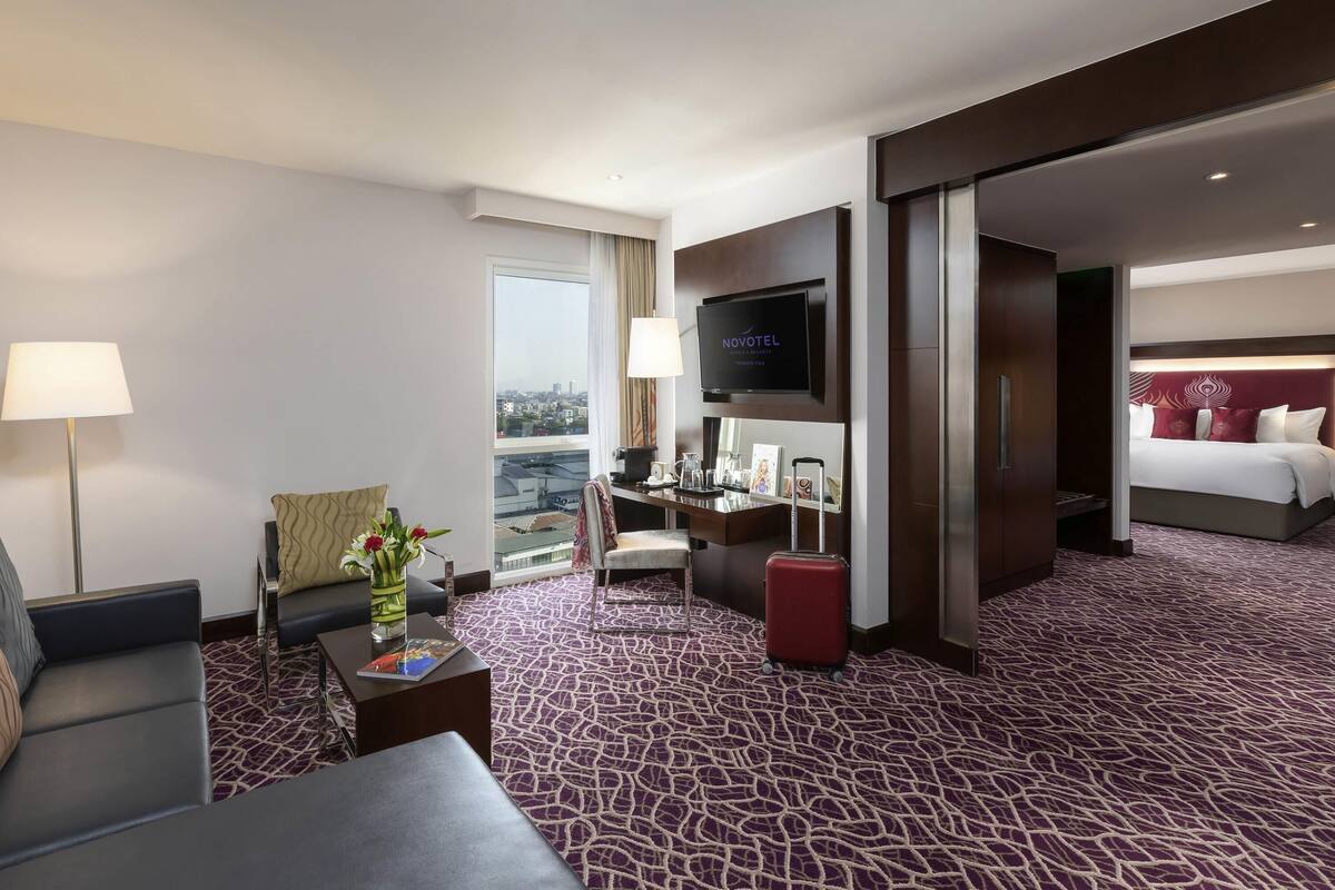 A hotel room with a large bed, a TV mounted on the wall, a desk with a chair, a sofa, a red suitcase, and a view of the city through the window.