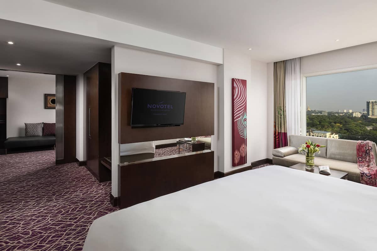A hotel room with a bed, a TV mounted on the wall, a sofa, and a large window offering a city view.