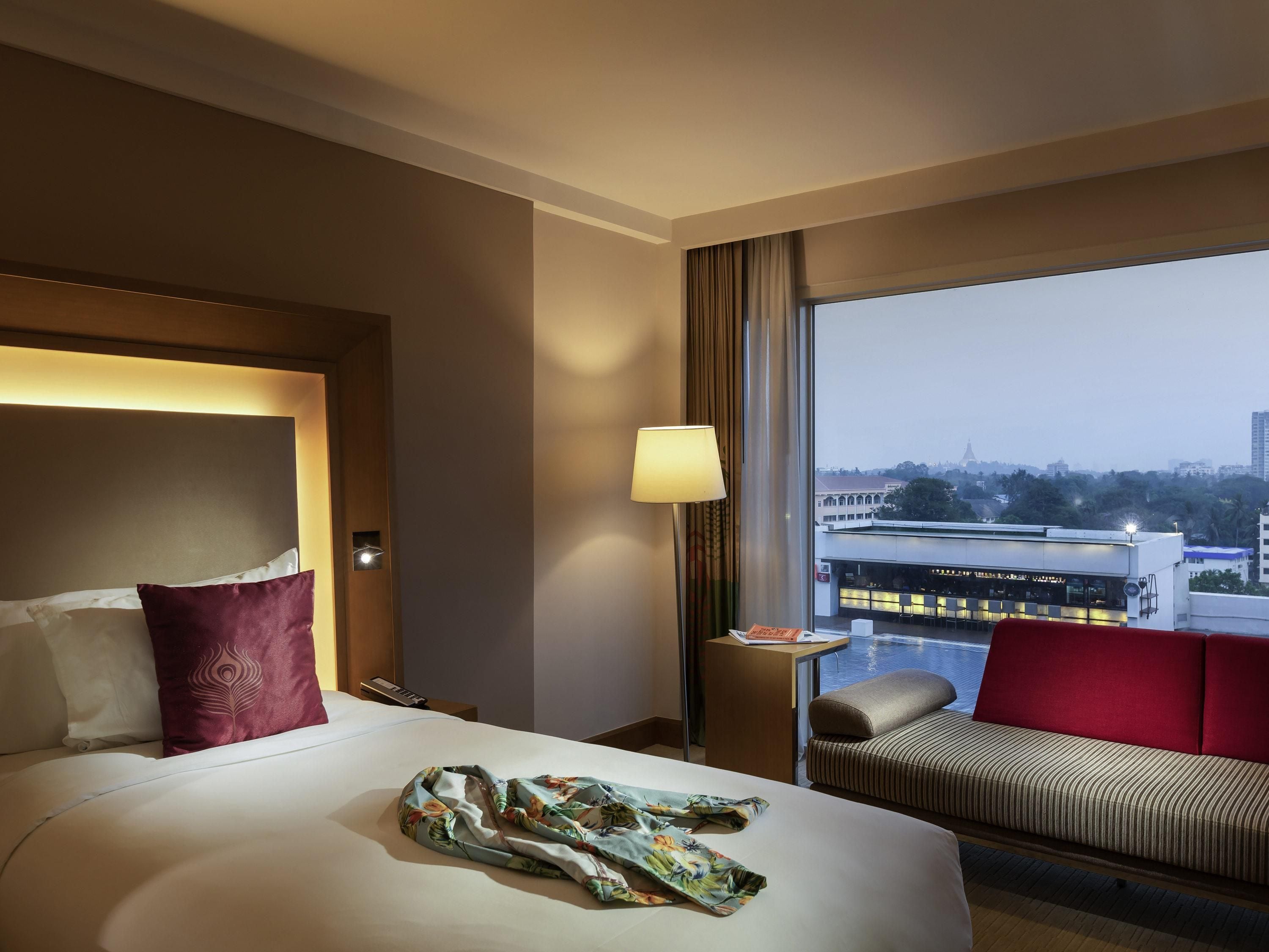 A hotel room with a bed, a sofa, a lamp, and a view of a cityscape.