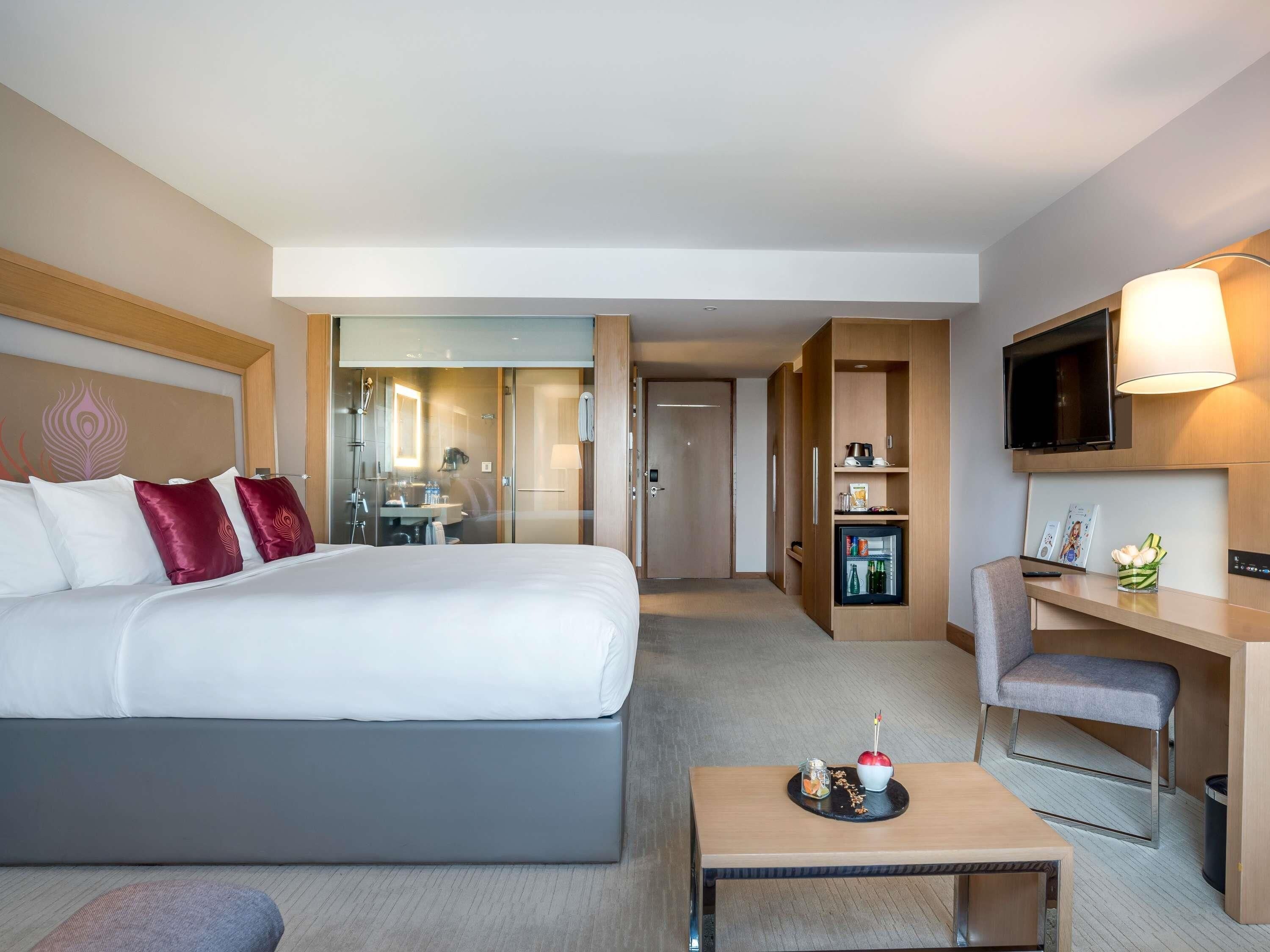 A hotel room with a bed, a desk, a TV, a mini-fridge, and a bathroom visible through a glass door.