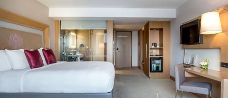 A hotel room with a bed, a desk, a TV, a mini-fridge, and a bathroom visible through a glass door.