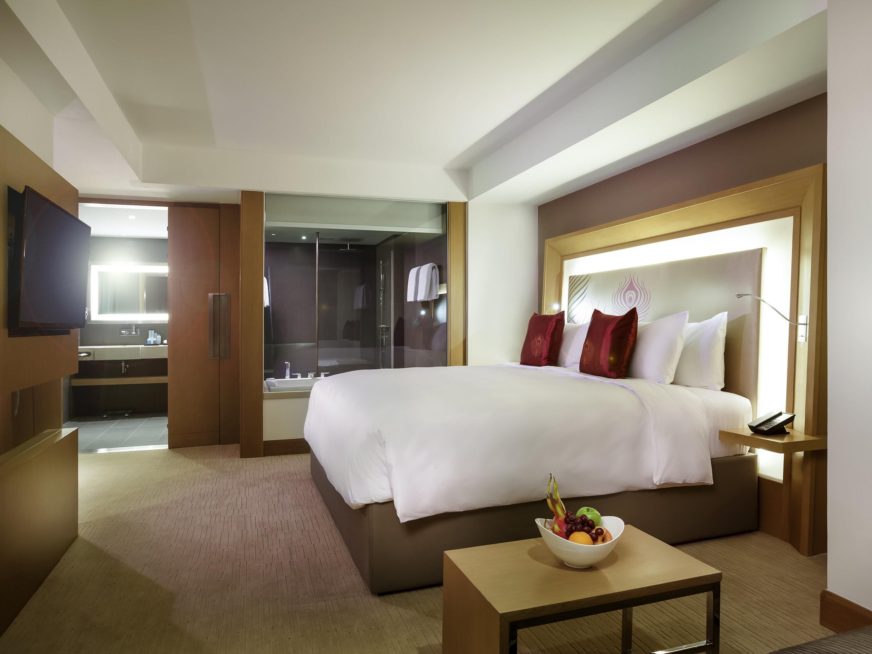 Habitación de hotel con una cama grande, una mesita de noche con un cuenco de fruta, un televisor, un baño con cabina de ducha de vidrio y un cabecero de madera.