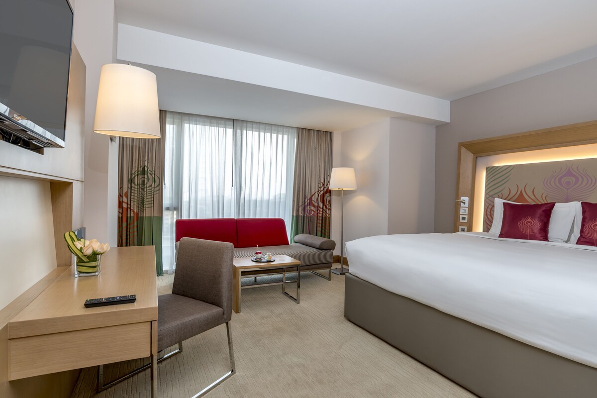 A hotel room with a bed, a desk with a remote control, a chair, a small table, and a sofa.