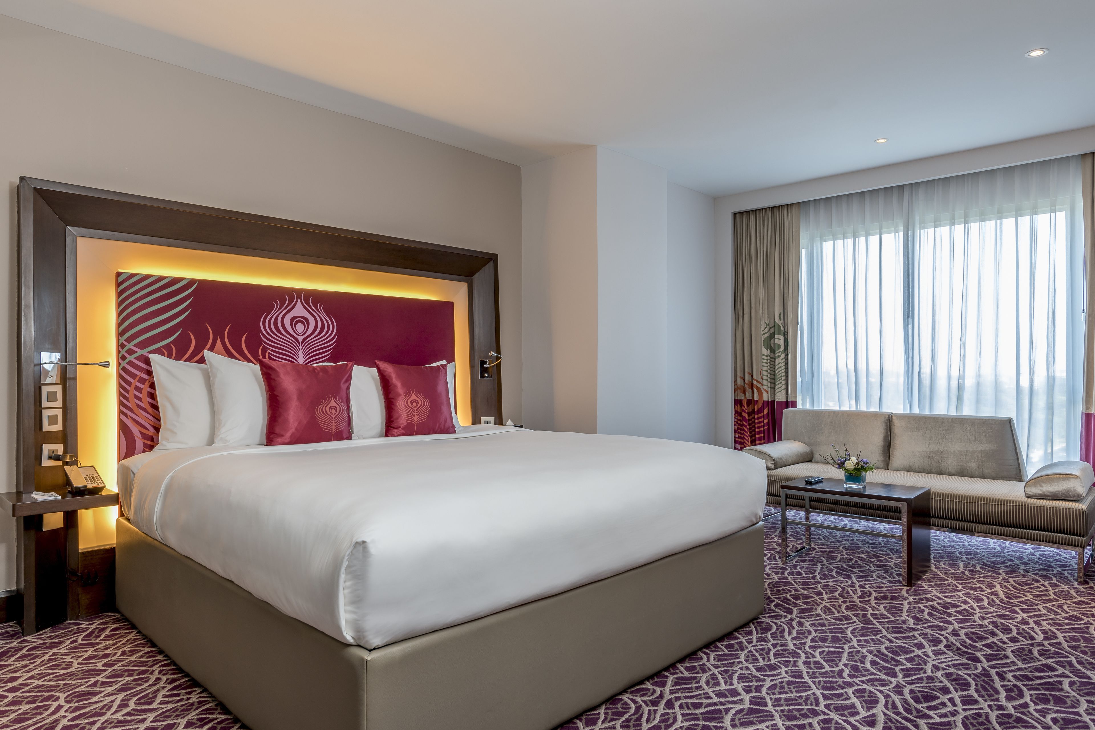 A hotel room with a large bed, a sofa, a coffee table, and a view of the city through large windows.