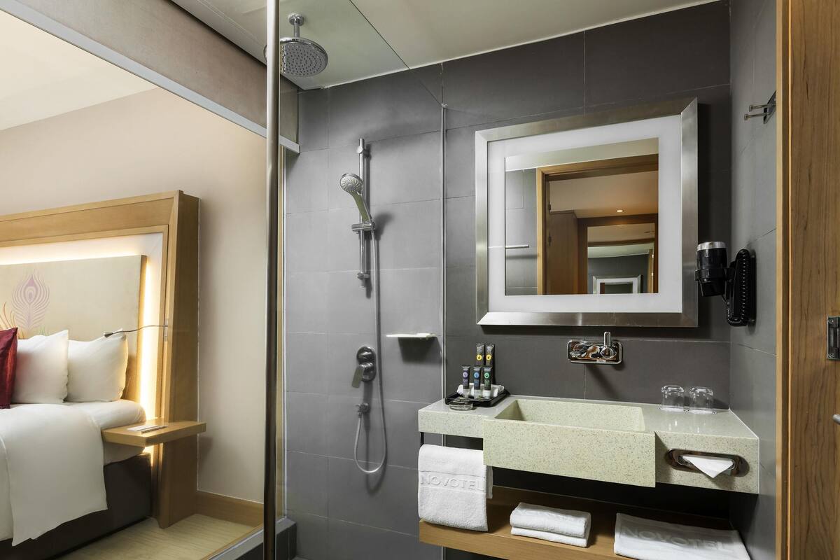 A hotel bathroom with a shower, a sink with toiletries, and a bed with a headboard.