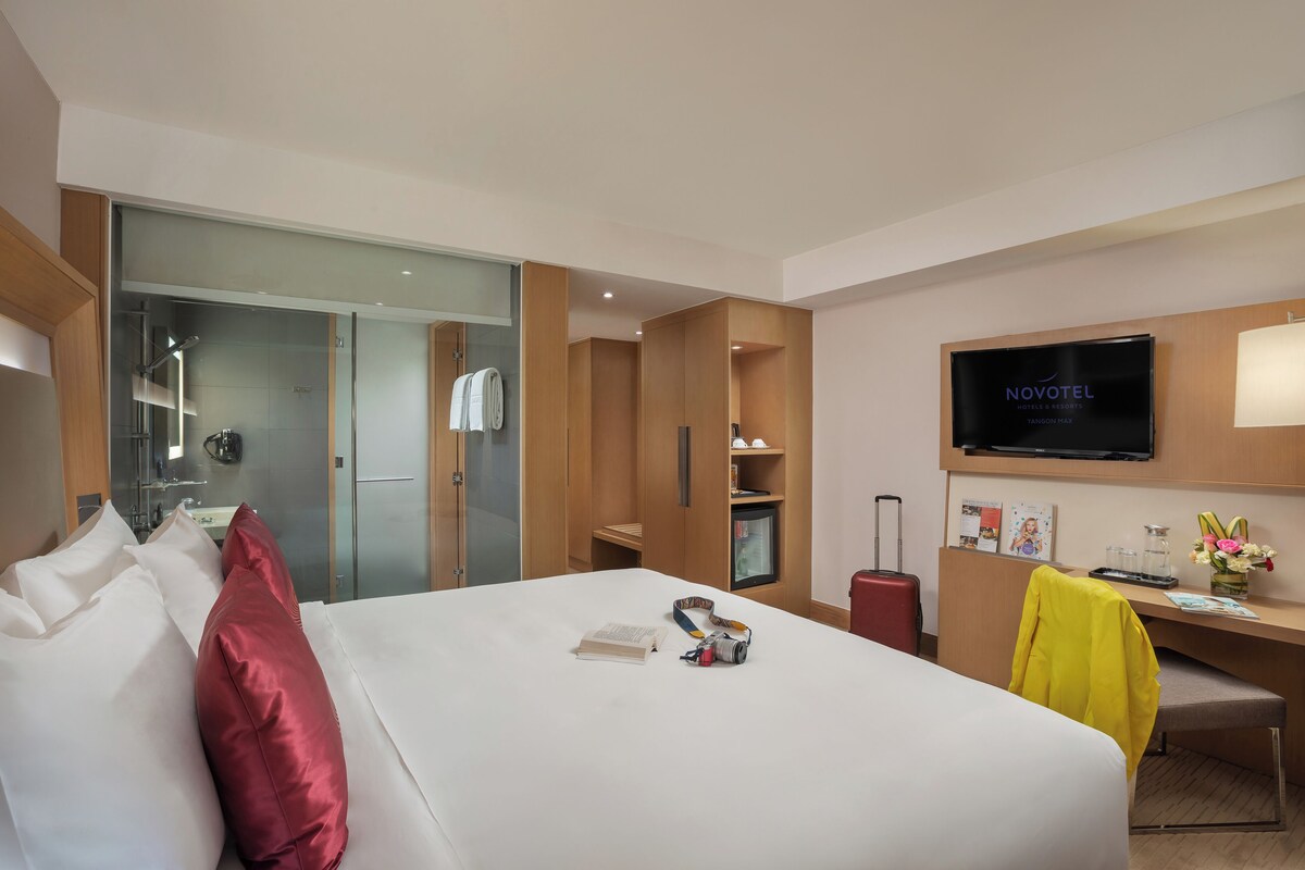 A hotel room with a bed, a desk, a TV, and a bathroom visible through a glass door.