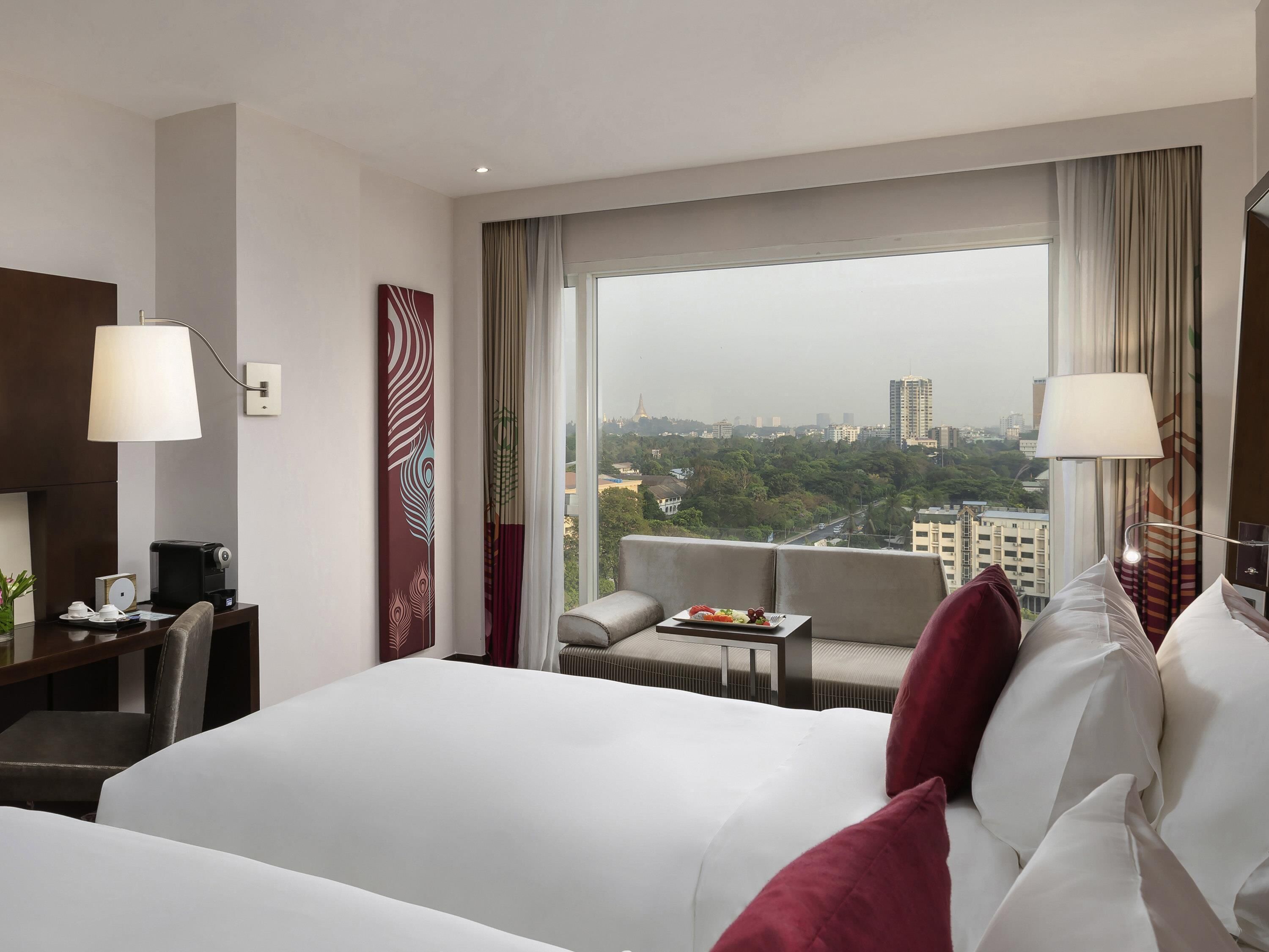 Habitación de hotel con cama, escritorio, silla, una mesita con bandeja de frutas y vistas a la ciudad a través de amplias ventanas.