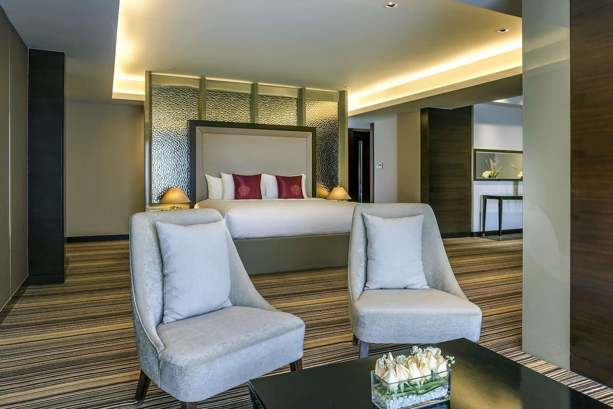 Une chambre d’hôtel avec un grand lit, deux fauteuils, une petite table ornée d’un vase de fleurs et un miroir au mur.