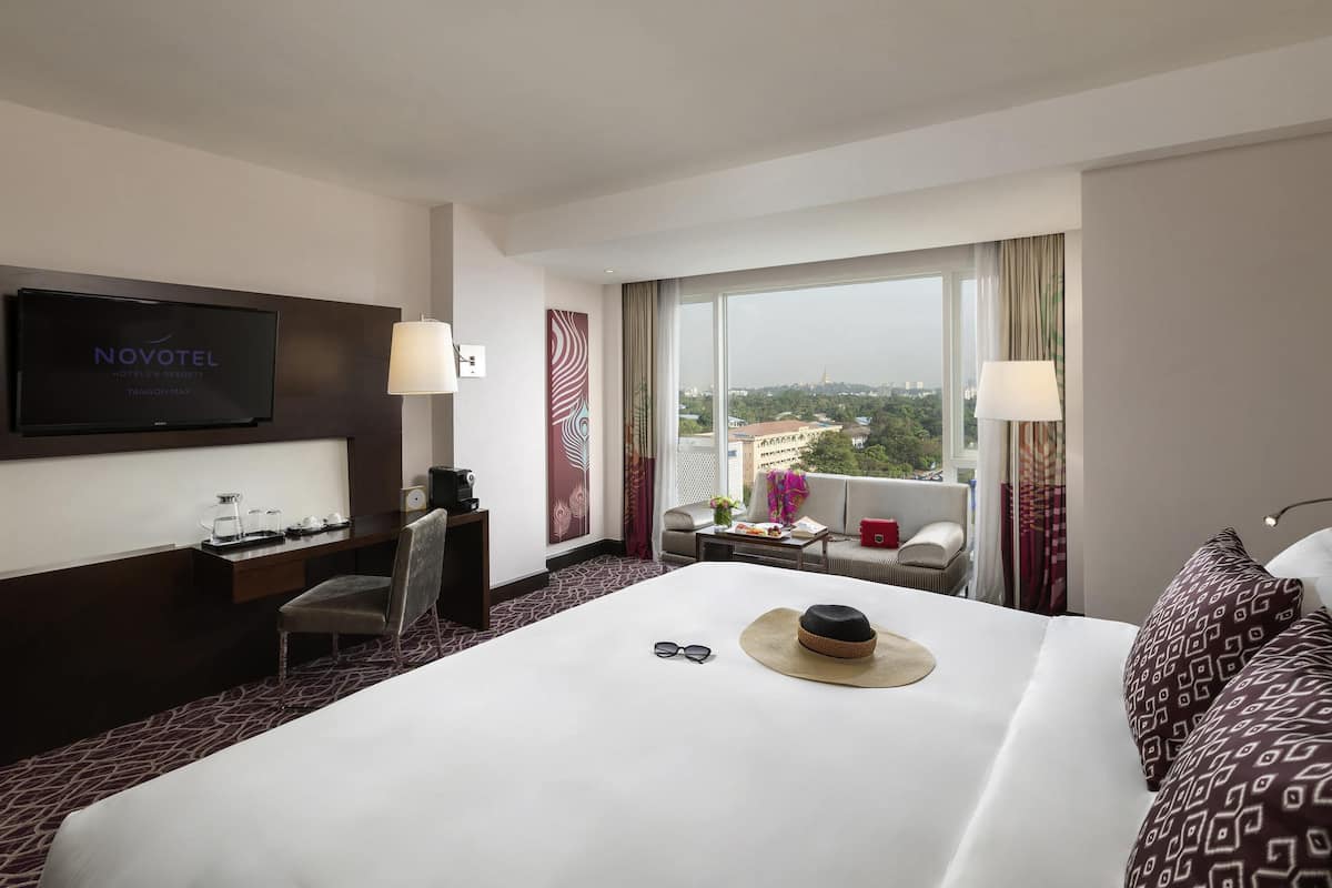 A hotel room with a bed, a TV, a desk with a chair, a window with a view, and a sofa.
