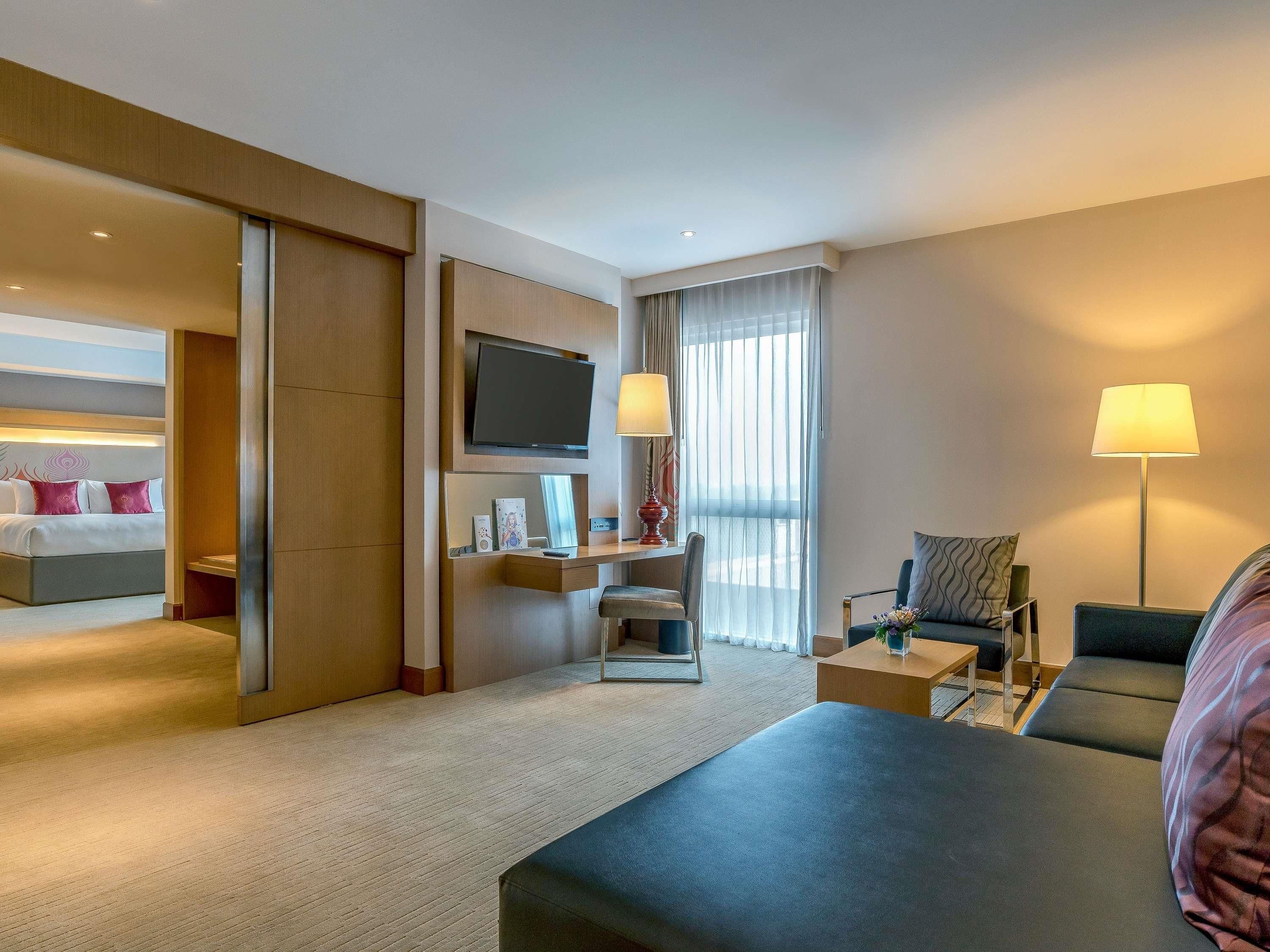 Quarto de hotel com cama, escrivaninha com televisão, cadeira, sofá e luminária.