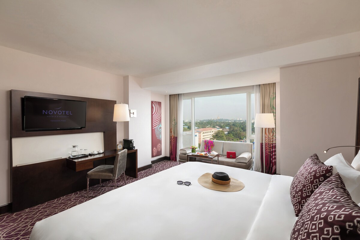 A hotel room with a bed, a desk, a TV, and a large window offering a city view.