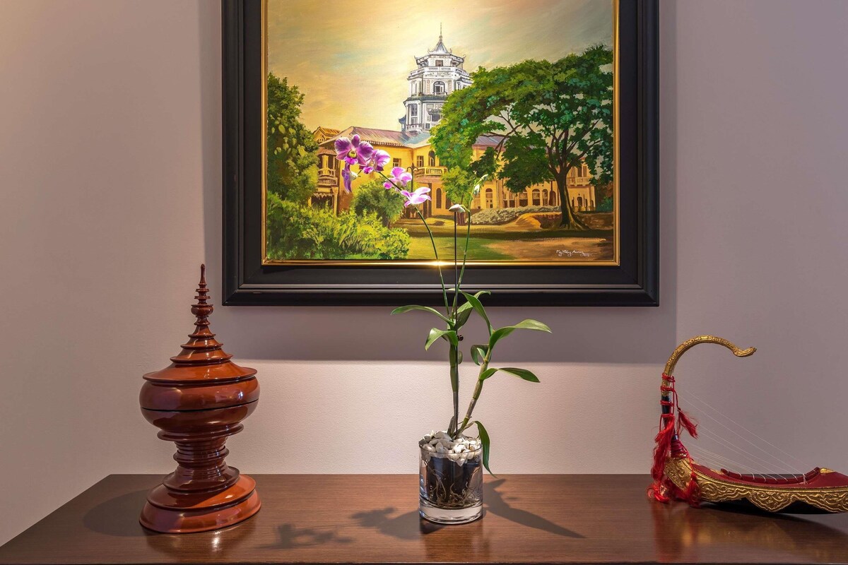 Un cuadro enmarco mostrando un edificio tradicional con una torre, un florero con una planta y un objeto decorativo sobre una mesa.