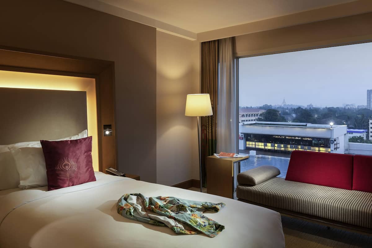 A hotel room with a bed, a sofa, a lamp, and a view of a cityscape.