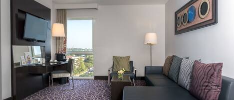 Habitación de hotel con una cama grande, un escritorio con computadora, un televisor montado en la pared y una ventana con vista a la ciudad.