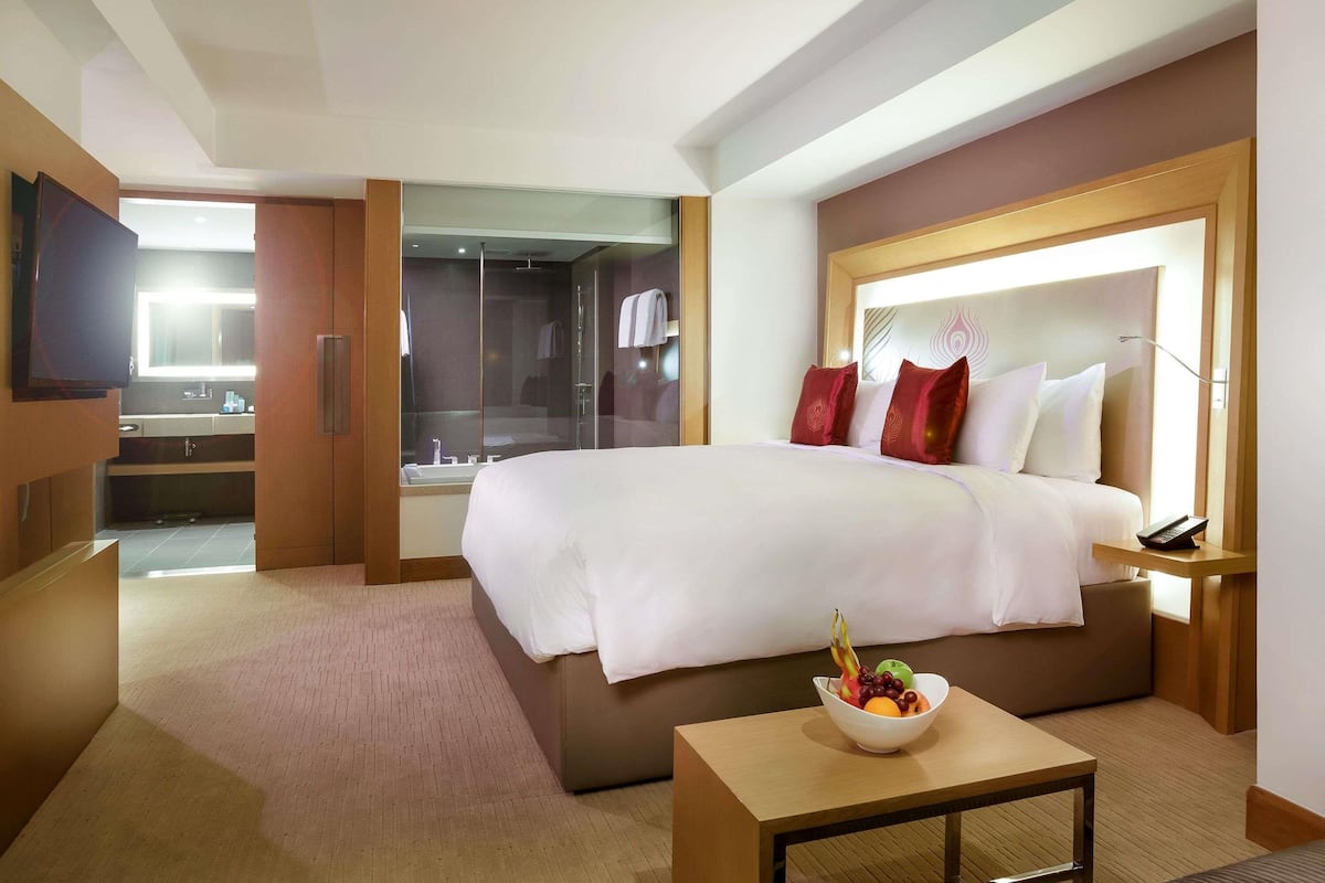 A hotel room with a large bed, a bedside table with a fruit bowl, a TV, a bathroom with a glass shower, and a wooden headboard.