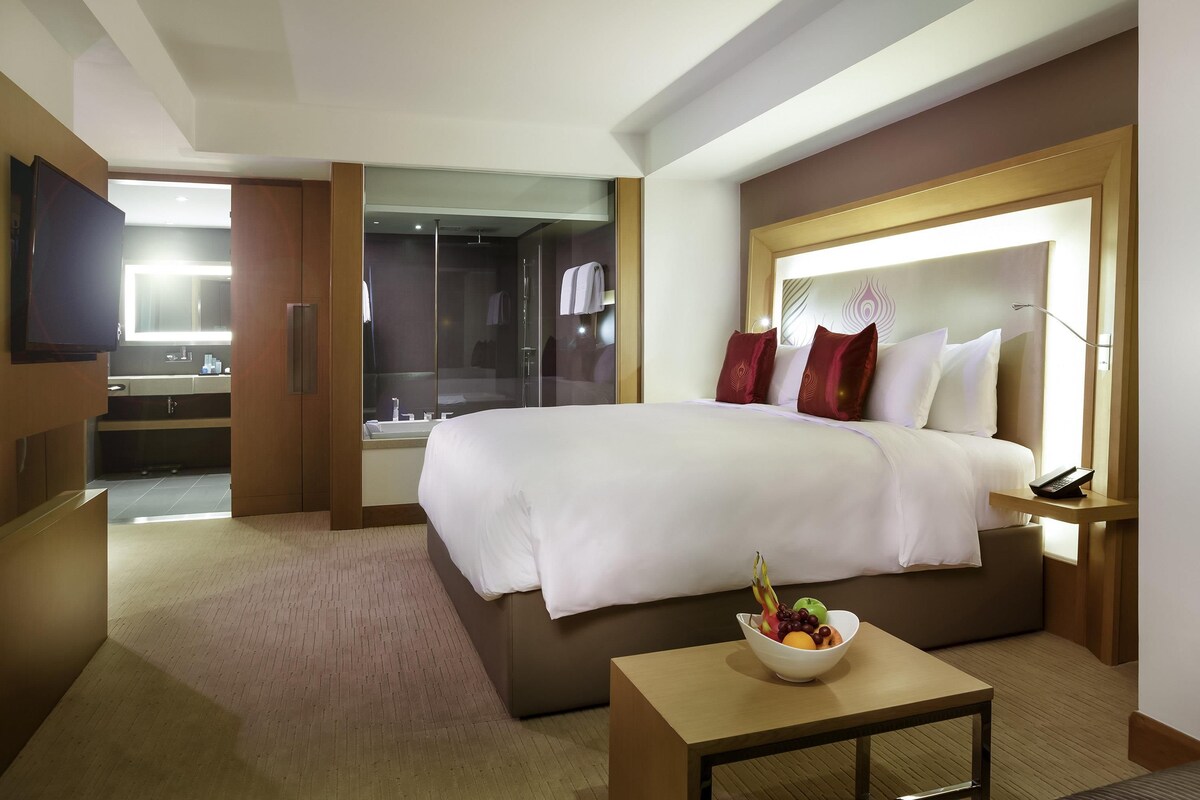 Habitación de hotel con una cama grande, una mesita de noche con un cuenco de fruta, un televisor, un baño con cabina de ducha de vidrio y un cabecero de madera.