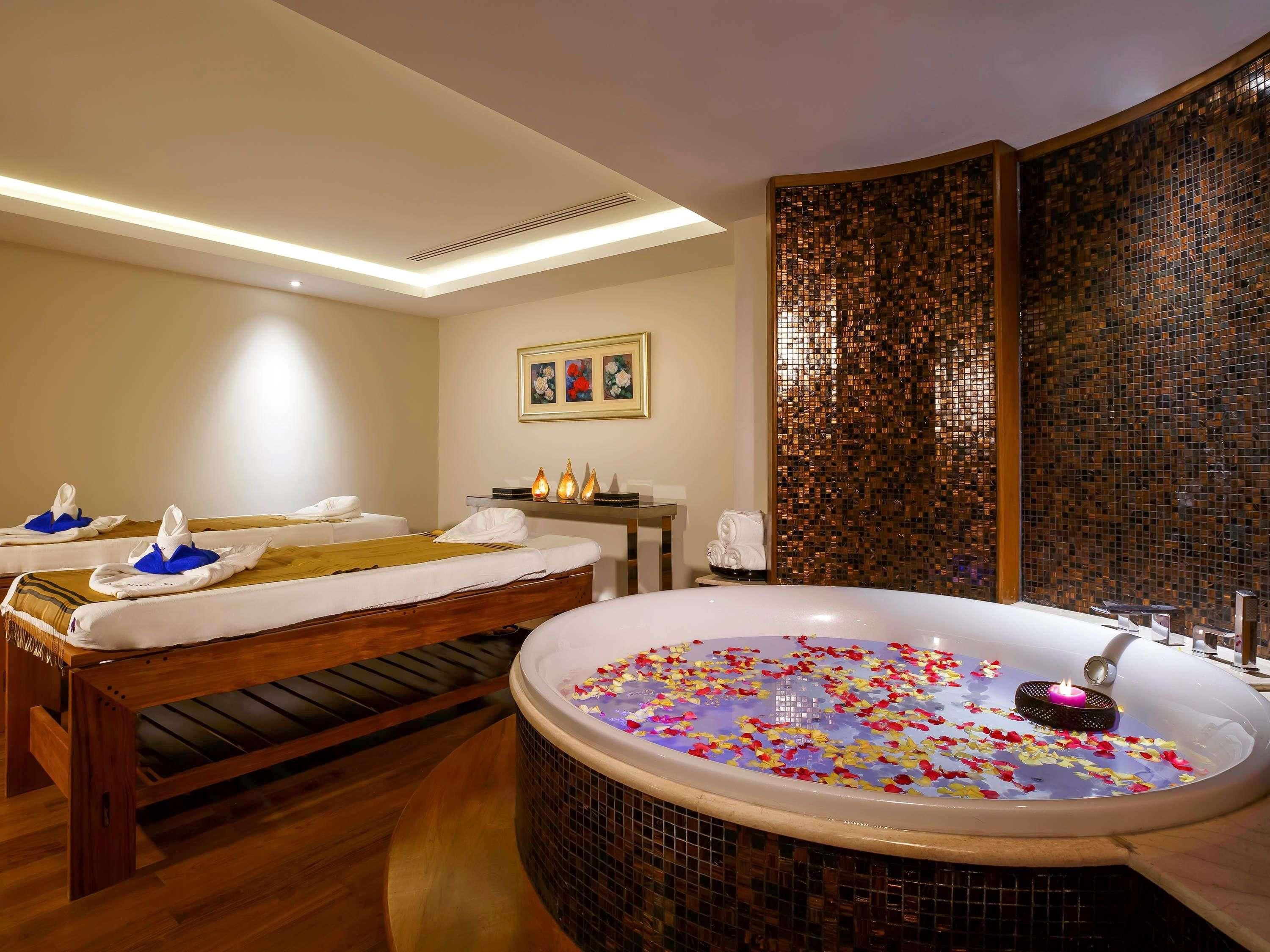 A spa room with two massage tables, a large jacuzzi with flower petals, and a decorative mosaic wall.