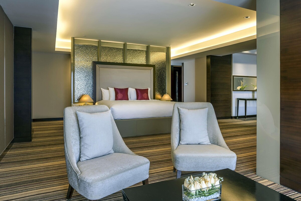 Una habitación de hotel con una cama grande, dos sillones, una mesita con un jarrón de flores y un espejo en la pared.