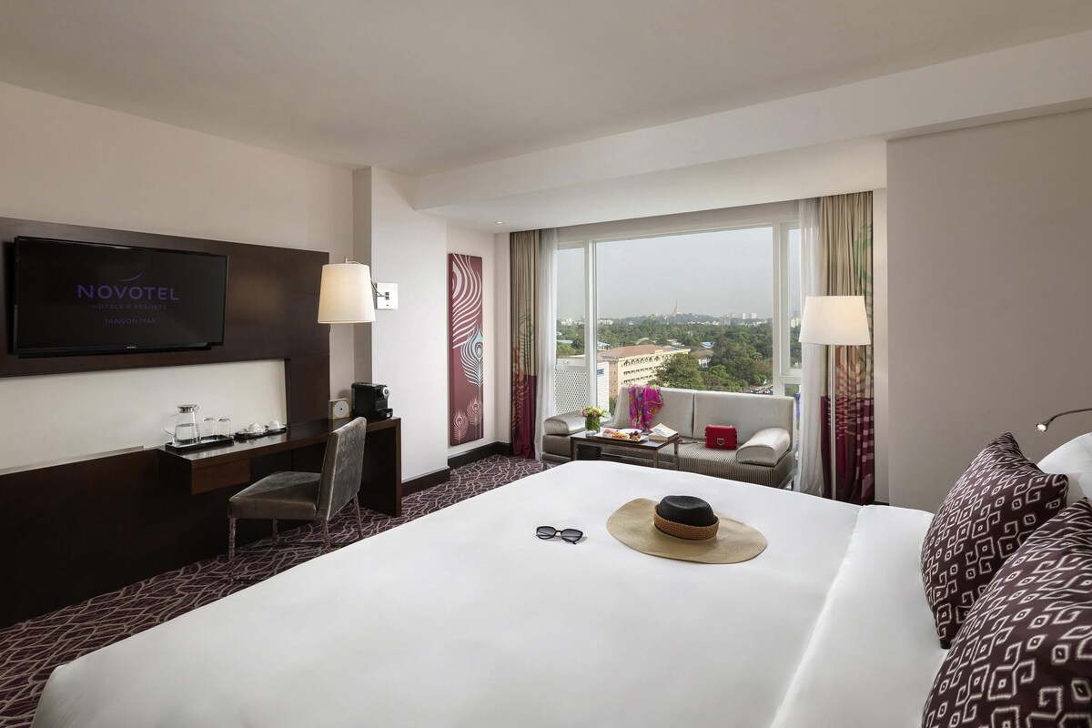 Habitación de hotel con cama, televisor, escritorio con silla, ventana con vistas y sofá.