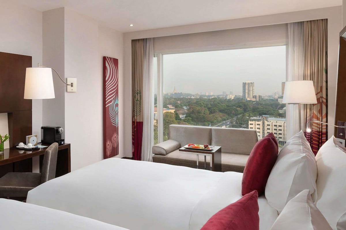 A hotel room with a large bed, a desk with a lamp, a small table with a plate of fruit, and a view of the city.