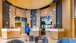 A modern hotel lobby with a reception desk, two receptionists, and a man on the phone. There are three blue chairs and a small table with a vase of flowers.