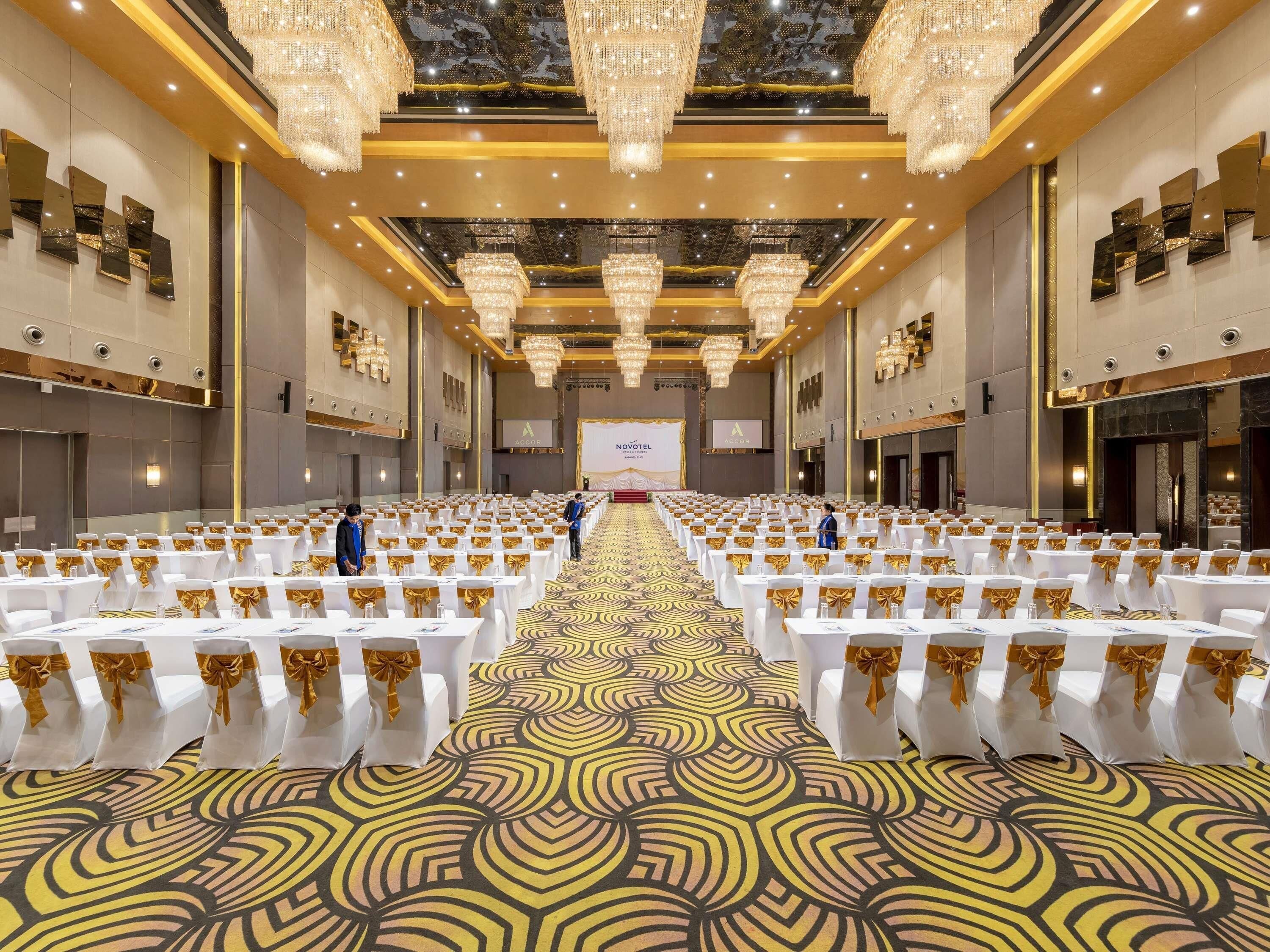 A spacious conference room with rows of white tables and chairs, gold sashes, and chandeliers.
