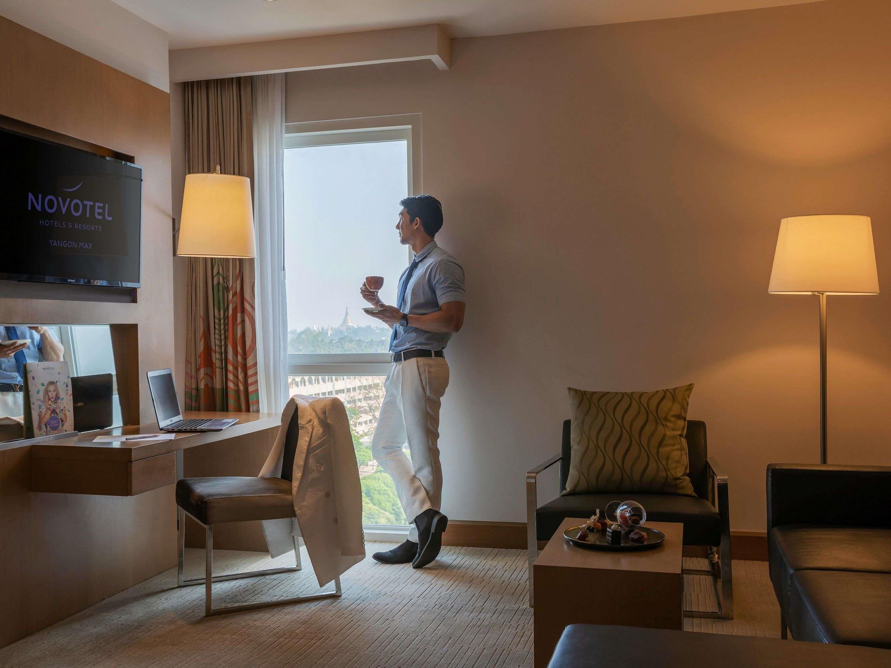 Habitación de hotel con televisor, escritorio, dos sillas, una mesa de centro y ventana con vistas.