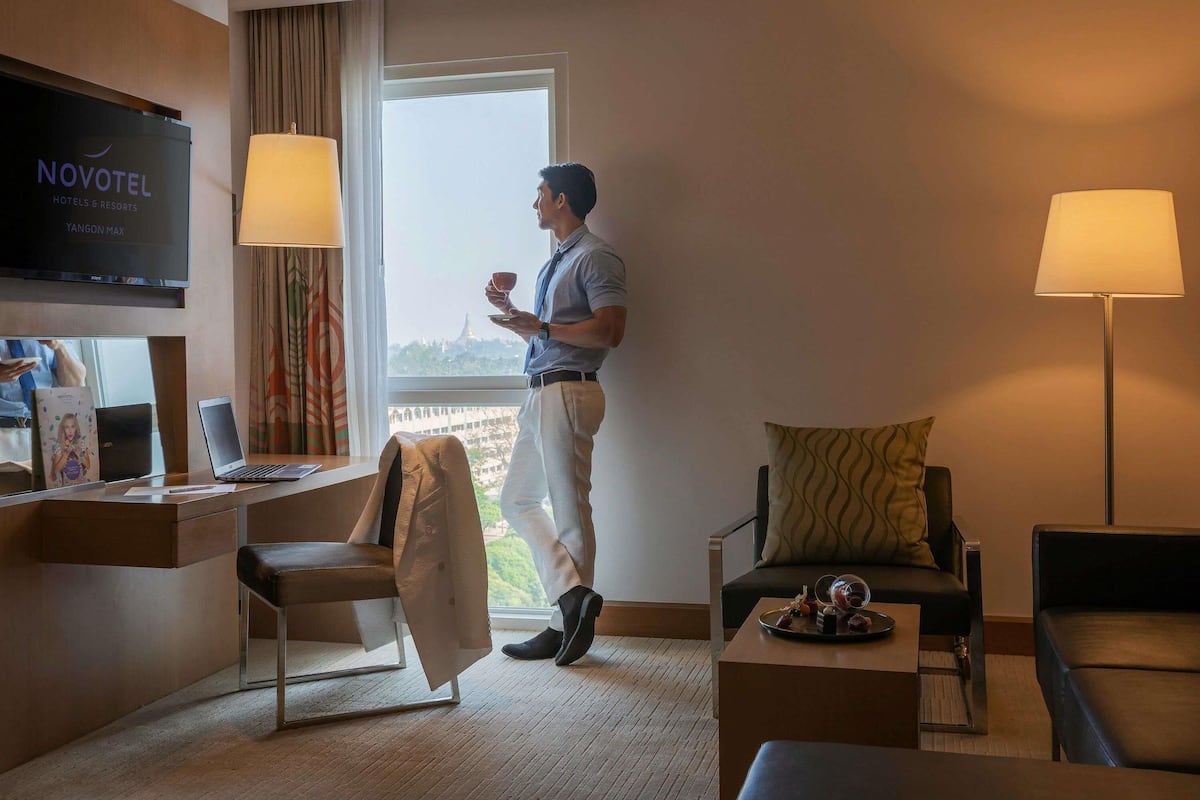 Habitación de hotel con televisor, escritorio, dos sillas, una mesa de centro y ventana con vistas.