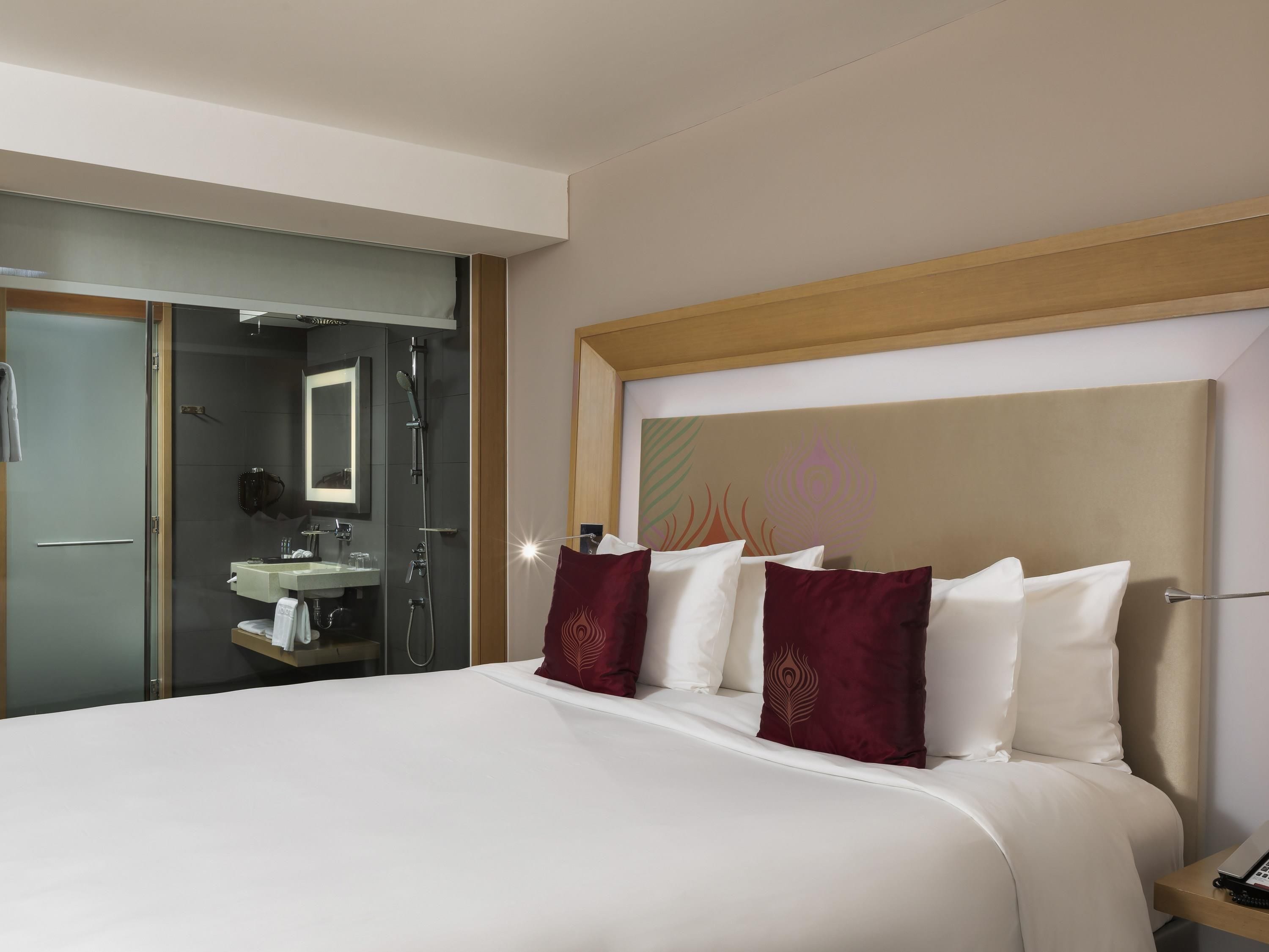 Una habitación de hotel con una cama grande, una mesita de noche con teléfono, un espejo, un baño con lavabo y ducha, y una pared con diseño decorativo.